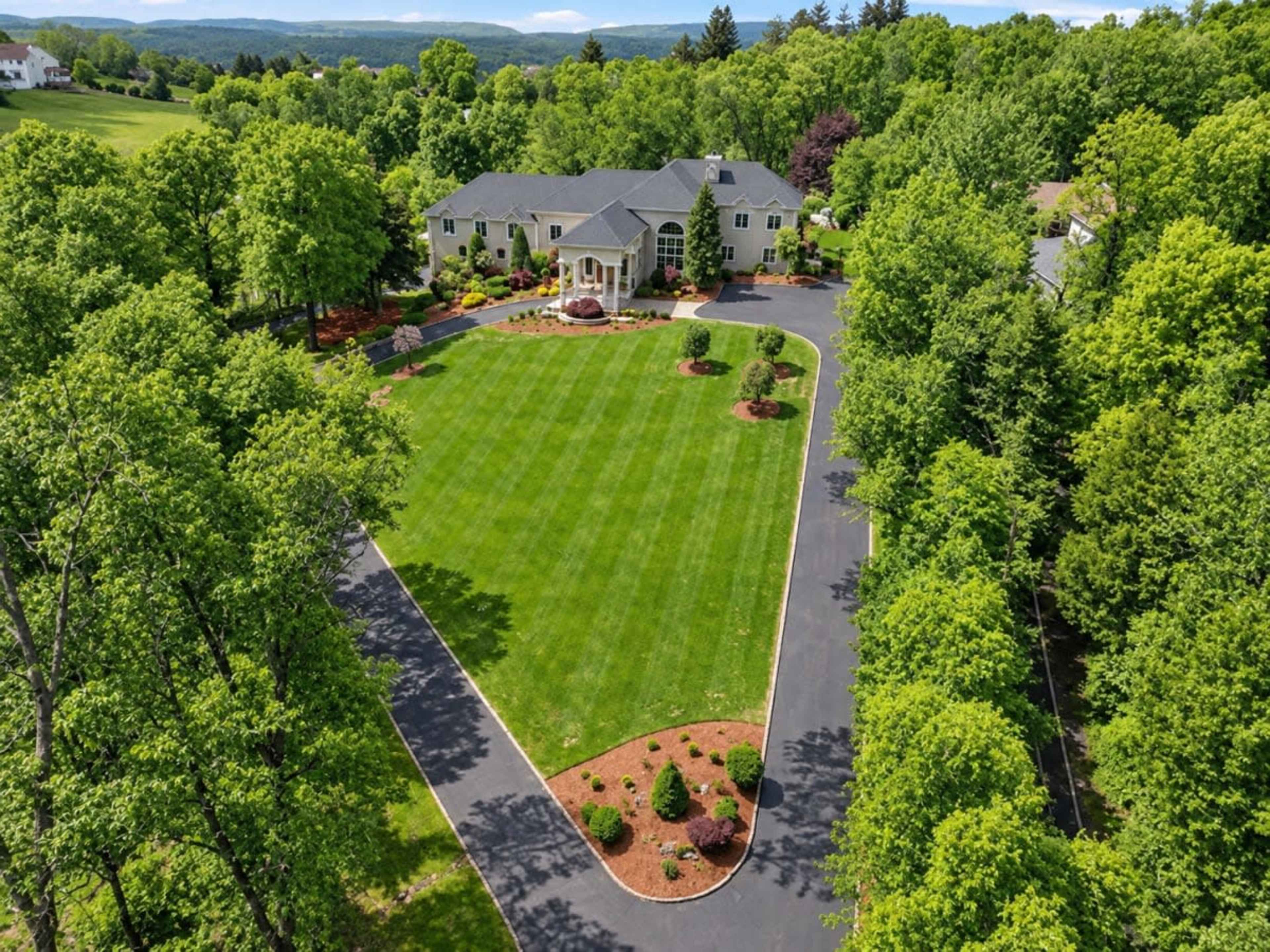 The image shows a large, two-story house surrounded by a landscaped lawn and trees, with a paved driveway leading to the entrance.