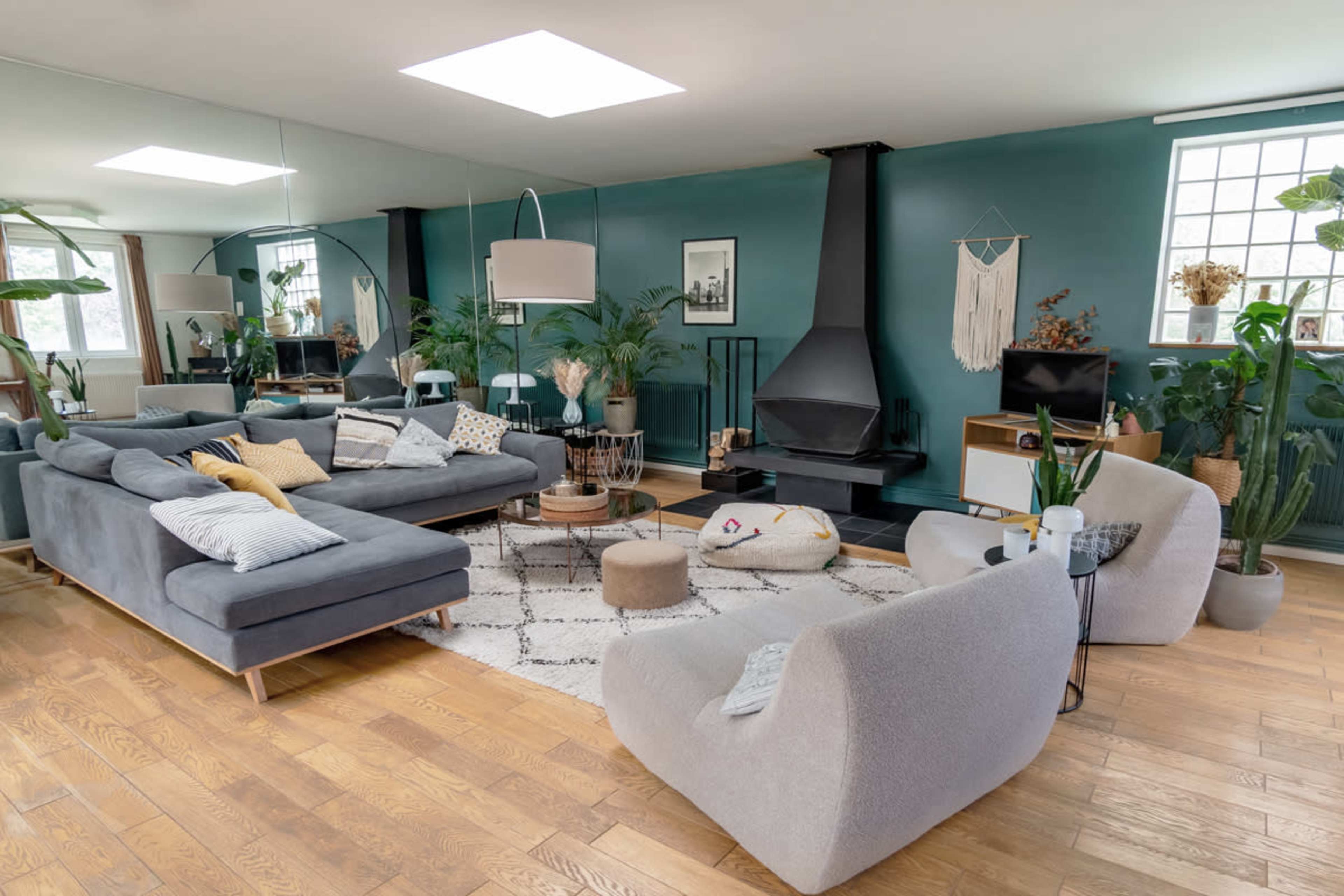 The image shows a modern living room with a gray sectional sofa, two light-colored armchairs, a wooden coffee table, and various indoor plants, all arranged around a central fireplace.