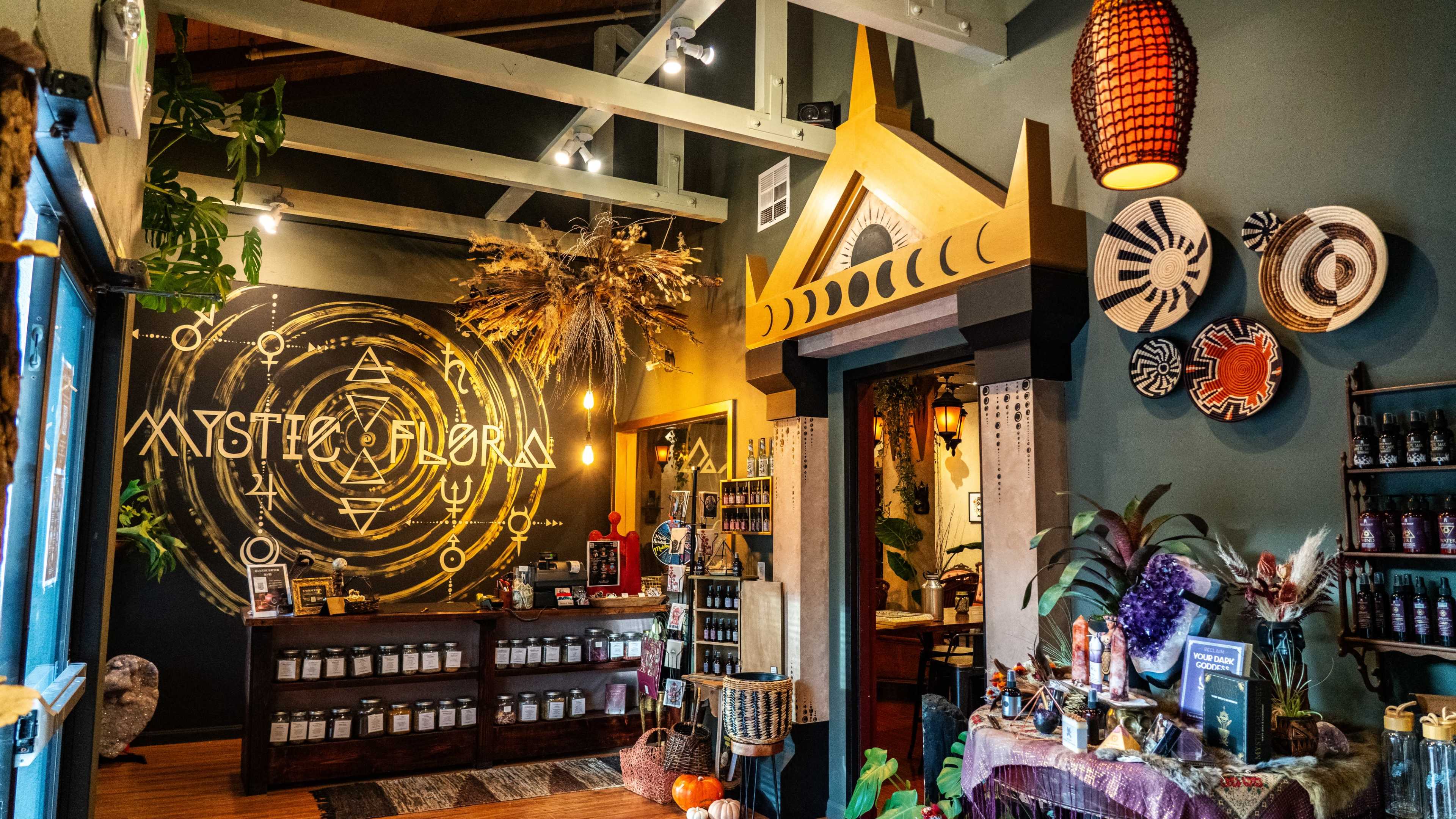 The image shows an interior of a shop named "Mystic Flora," featuring decorative elements, shelves with jars, and a wall adorned with a circular design.