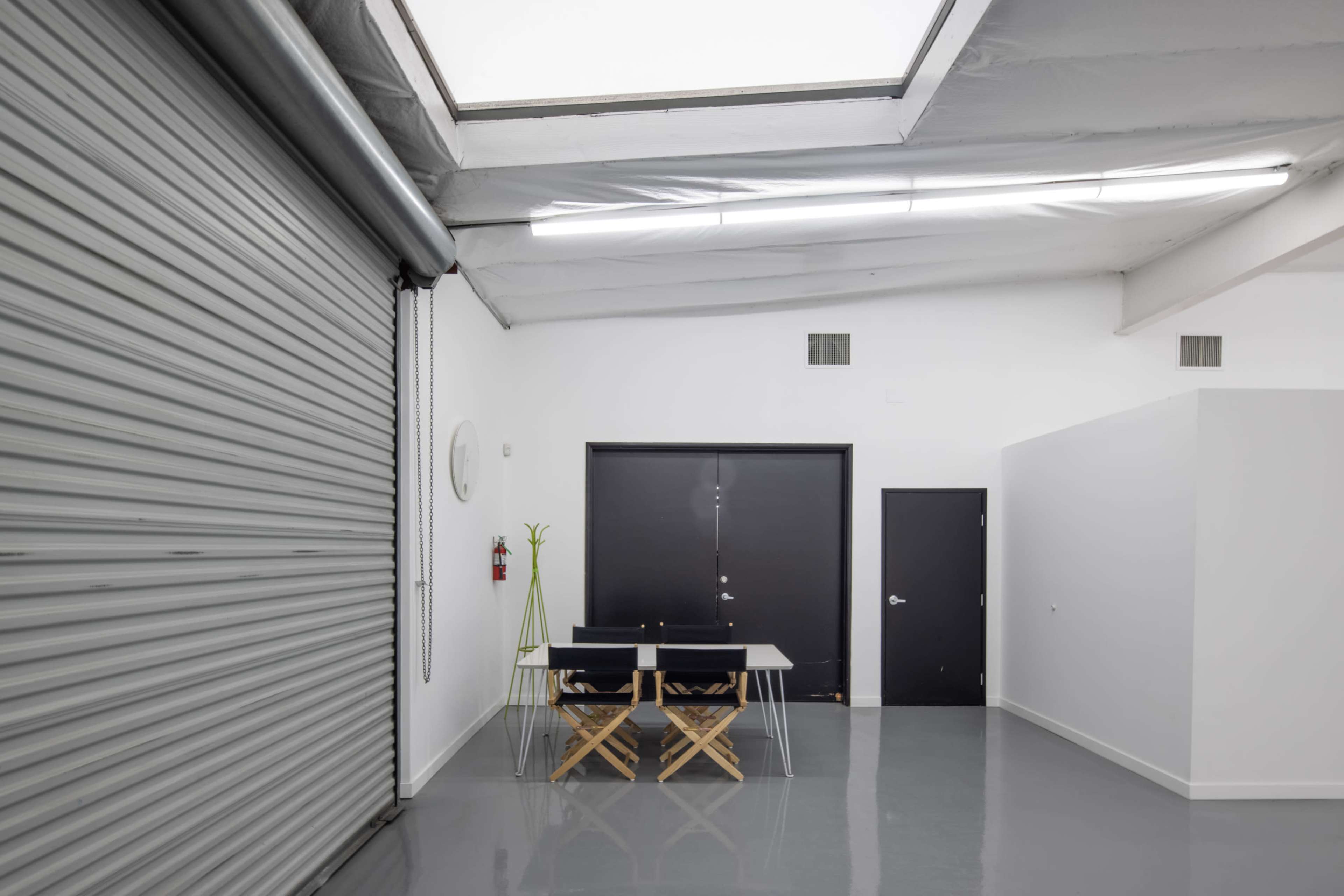 A minimalist interior features a folding table with chairs next to a closed metal roll-up door and a single door framed by white walls and a skylight.