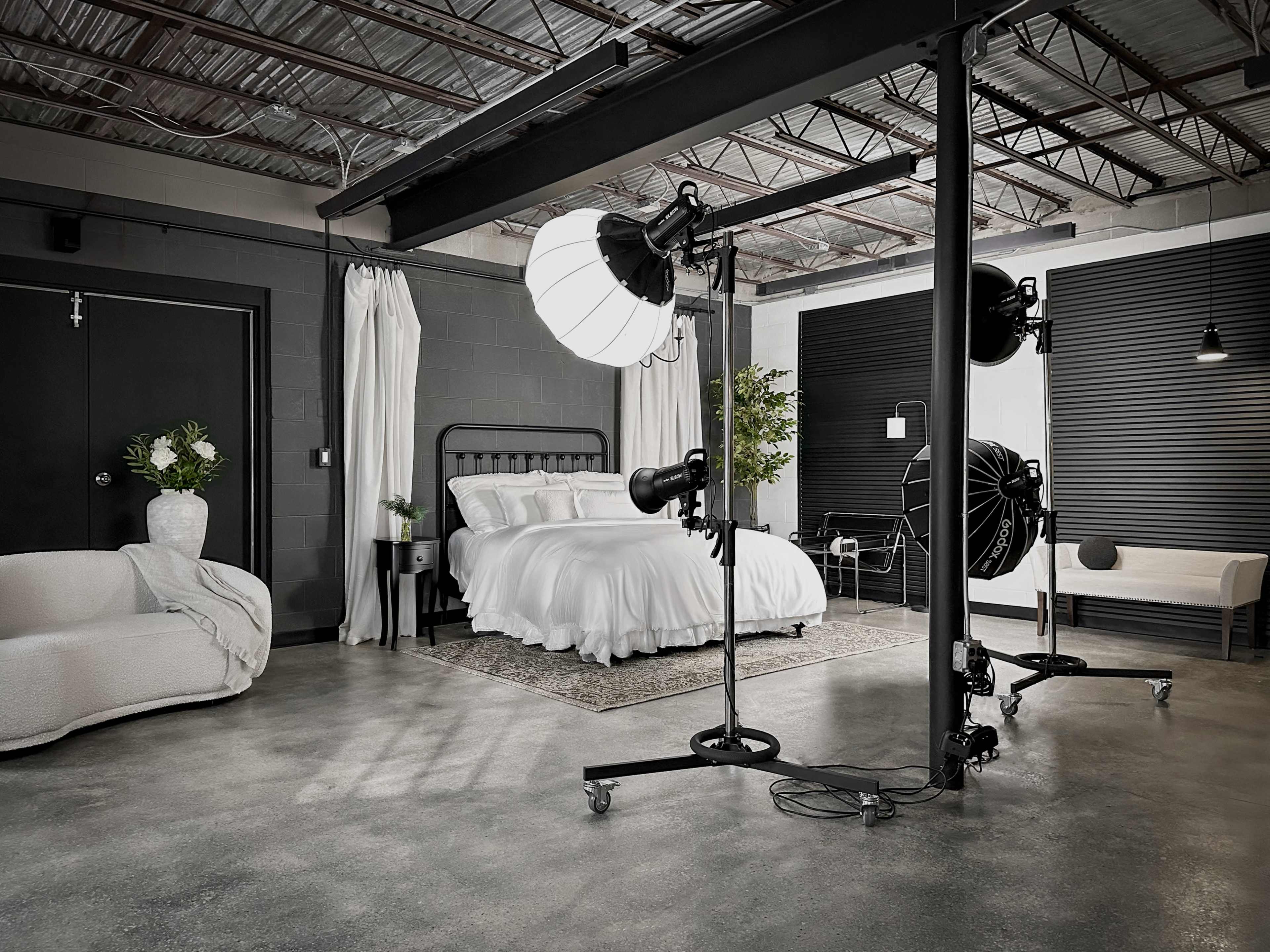 The image shows a modern studio space featuring a well-lit bedroom with a bed covered in white linens, surrounded by decorative plants and various studio lighting equipment.
