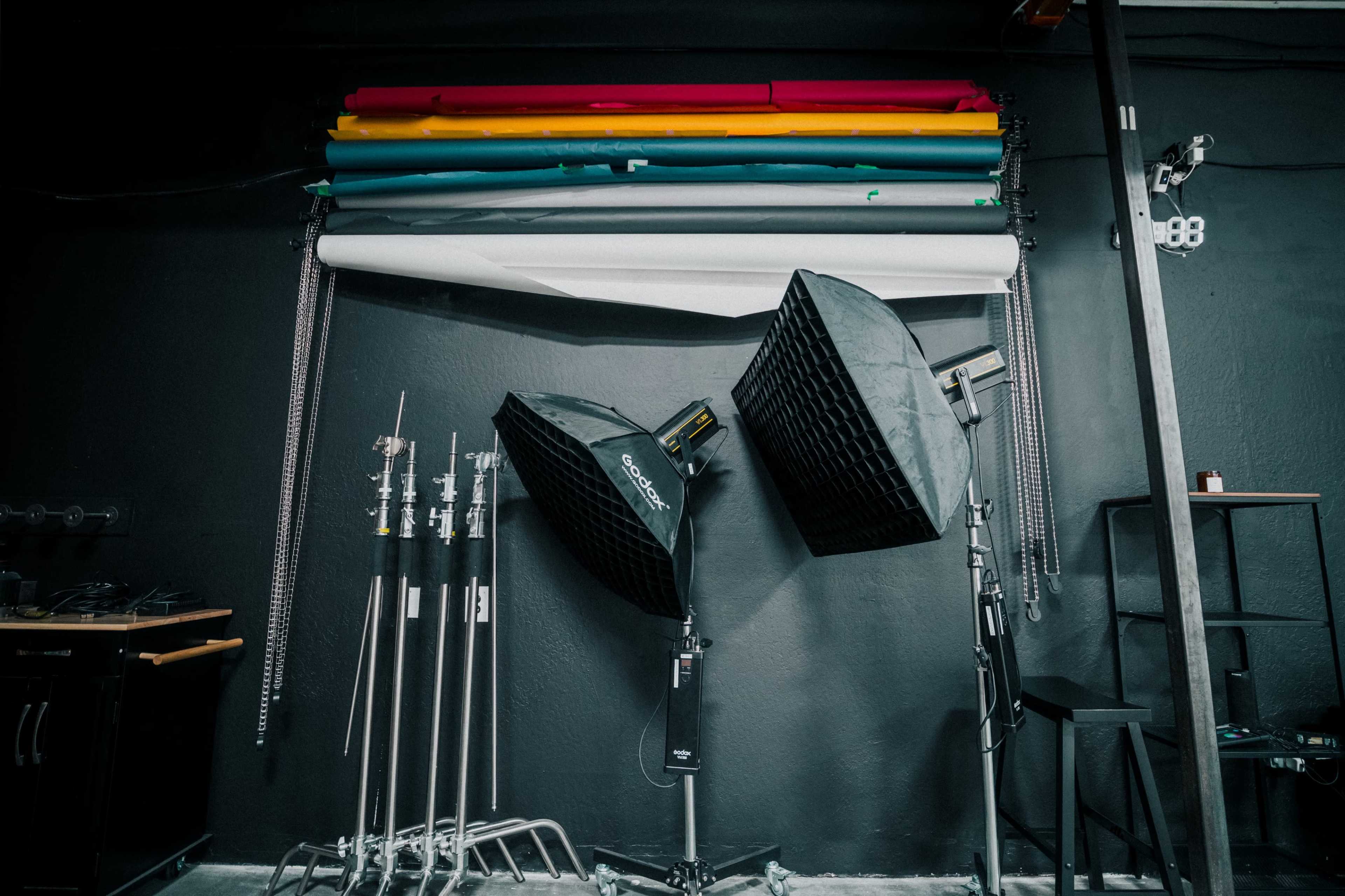 The image shows a photography studio with backdrop rolls in various colors hanging on the wall and two softbox lights positioned on stands.