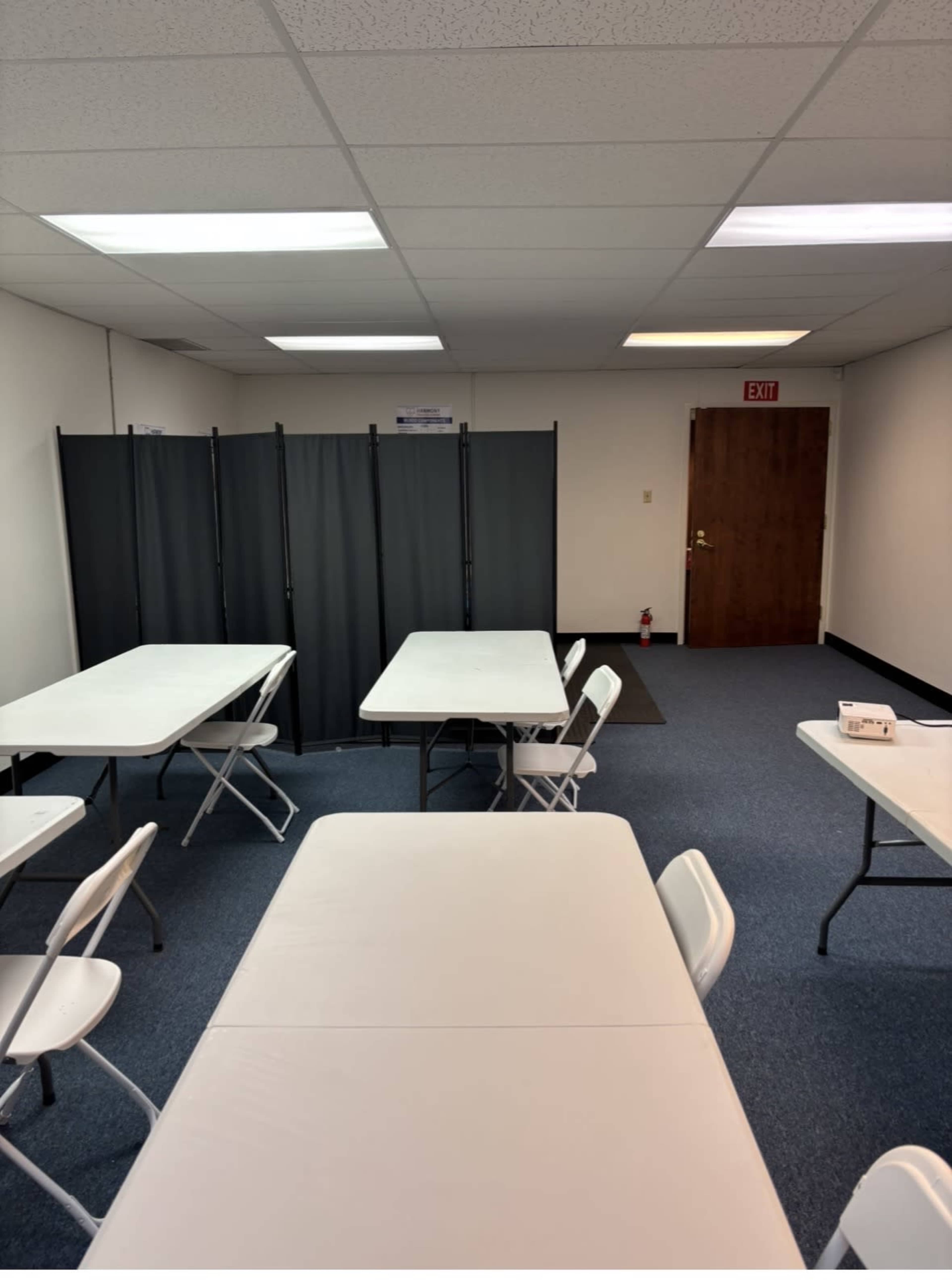 The image shows a sparsely furnished room with several white folding tables and chairs arranged, a gray partition in the background, and a wooden door leading to another space.