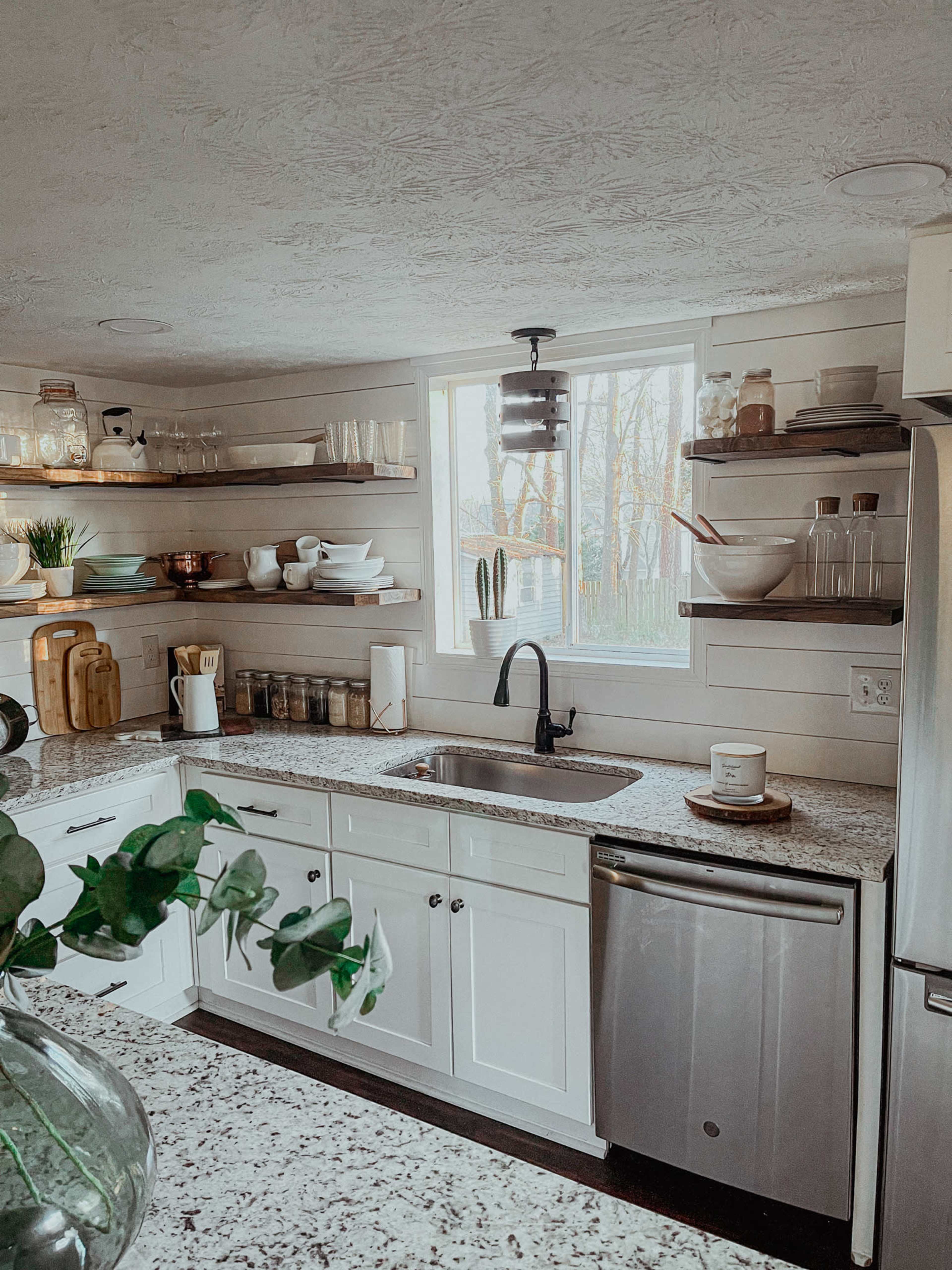 A modern kitchen features white cabinetry, open shelves with various dishware, and a window overlooking greenery.