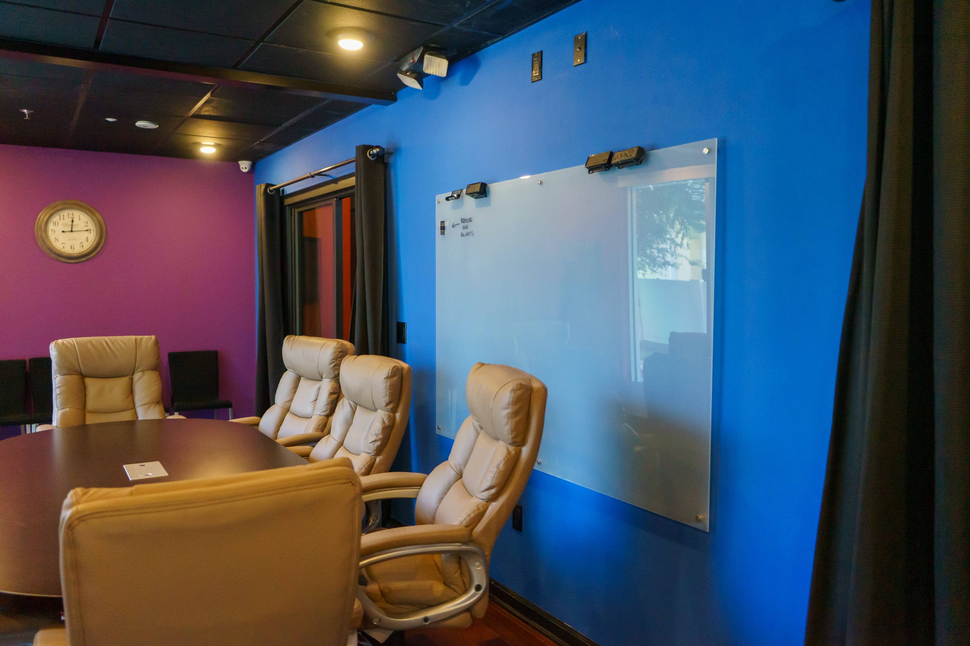 A conference room features a round table surrounded by light-colored chairs, a whiteboard on the blue wall, and a purple accent wall.