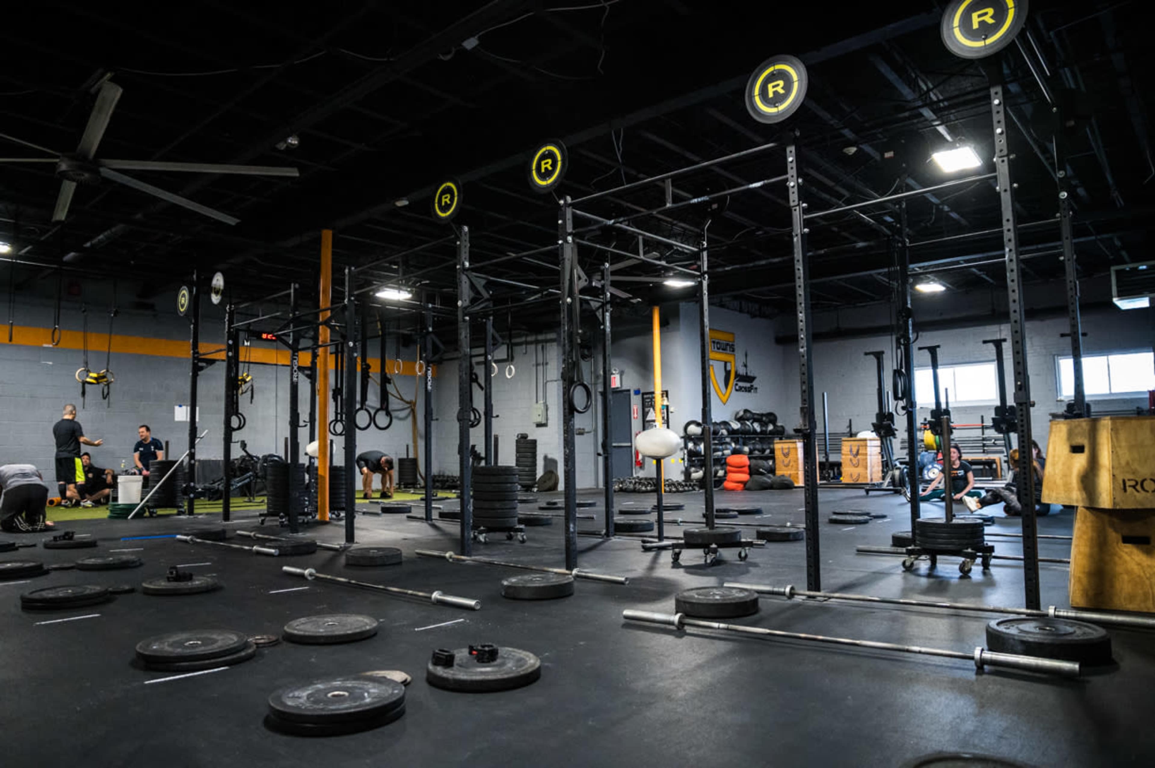 The image shows a fitness gym with multiple weightlifting stations, benches, and numerous weight plates scattered across the floor.