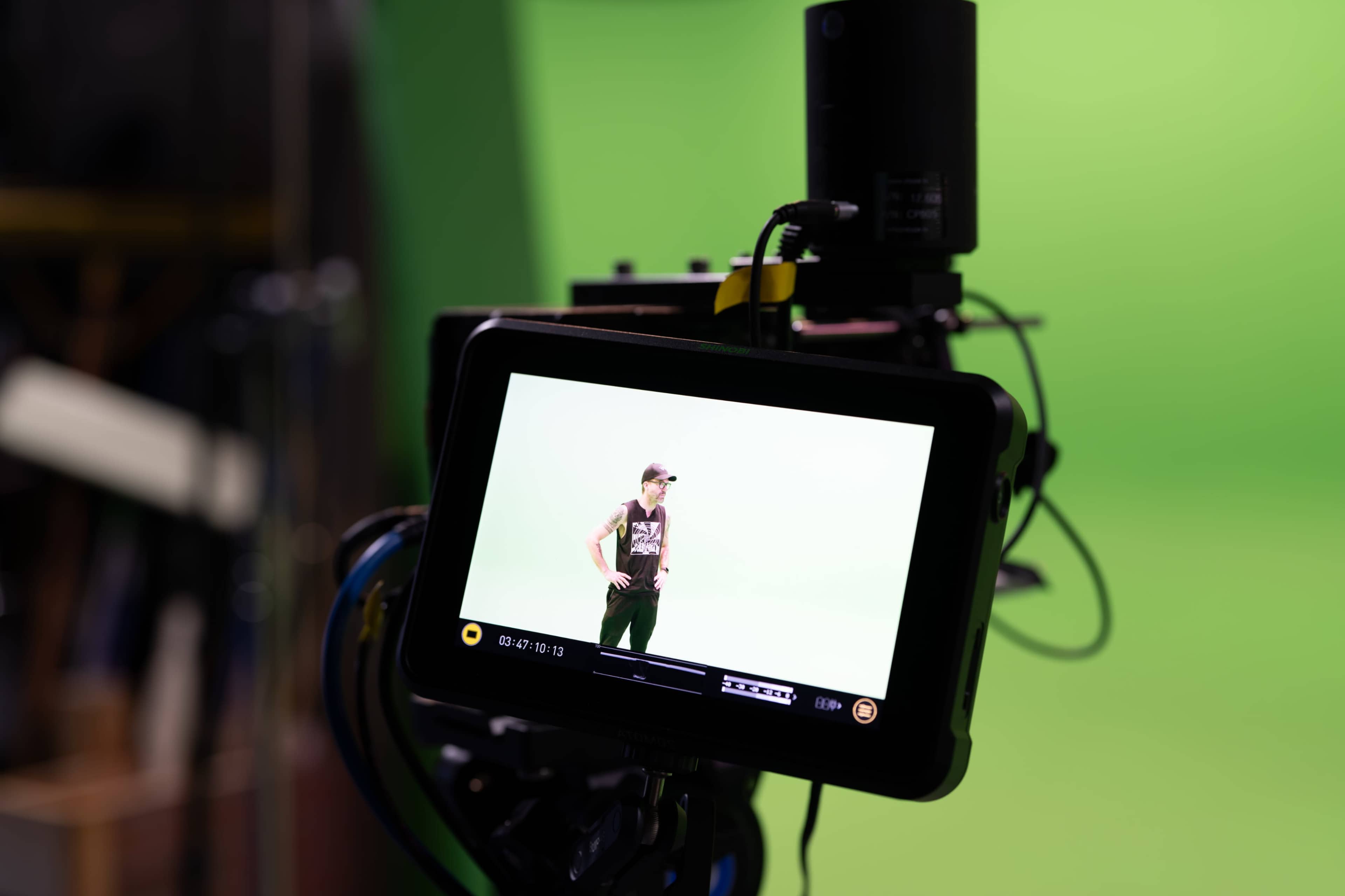 A camera monitor displays a person posing in front of a green screen.