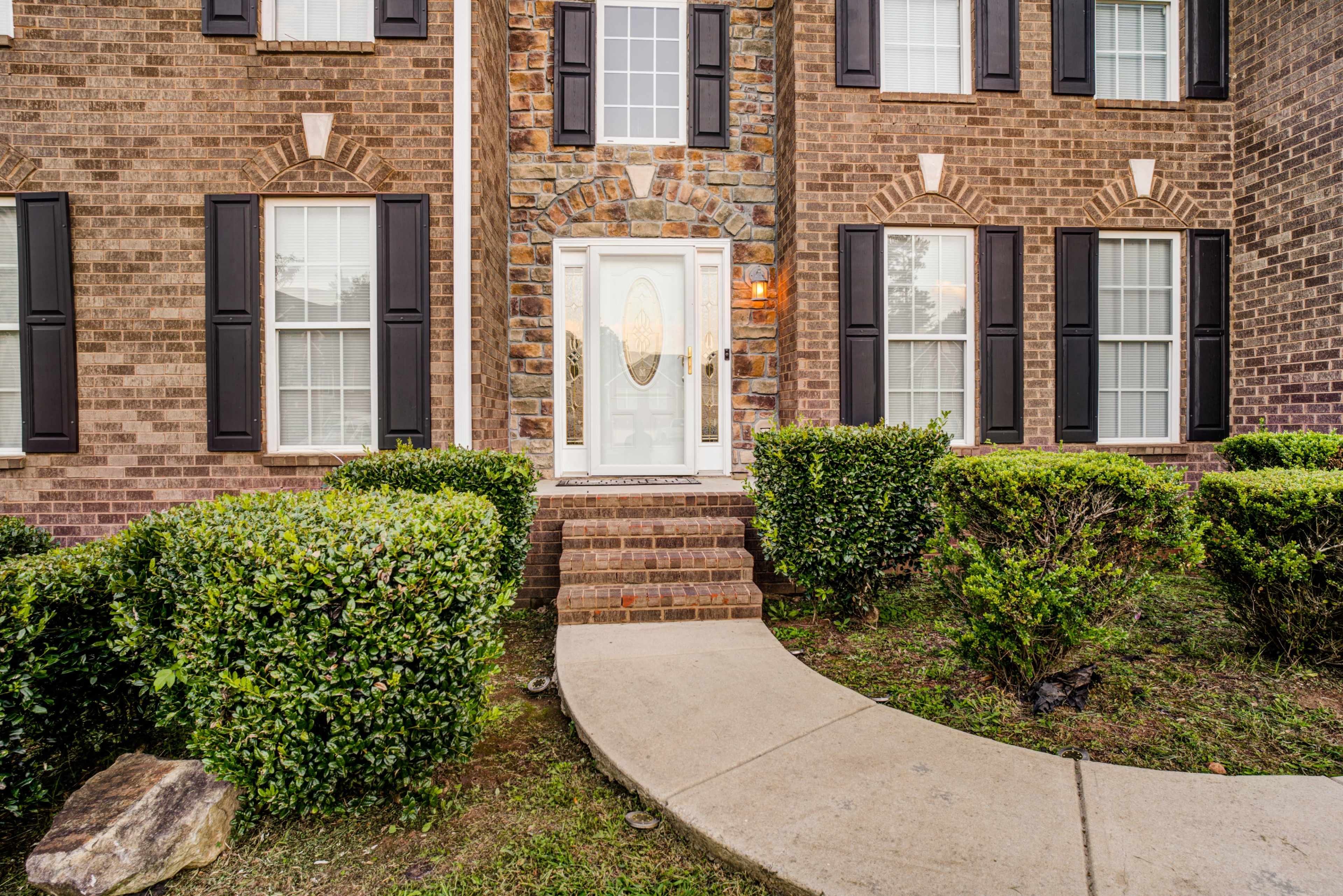 The entrance of a brick house with a stone-adorned door surrounded by neatly trimmed bushes and a curved walkway.