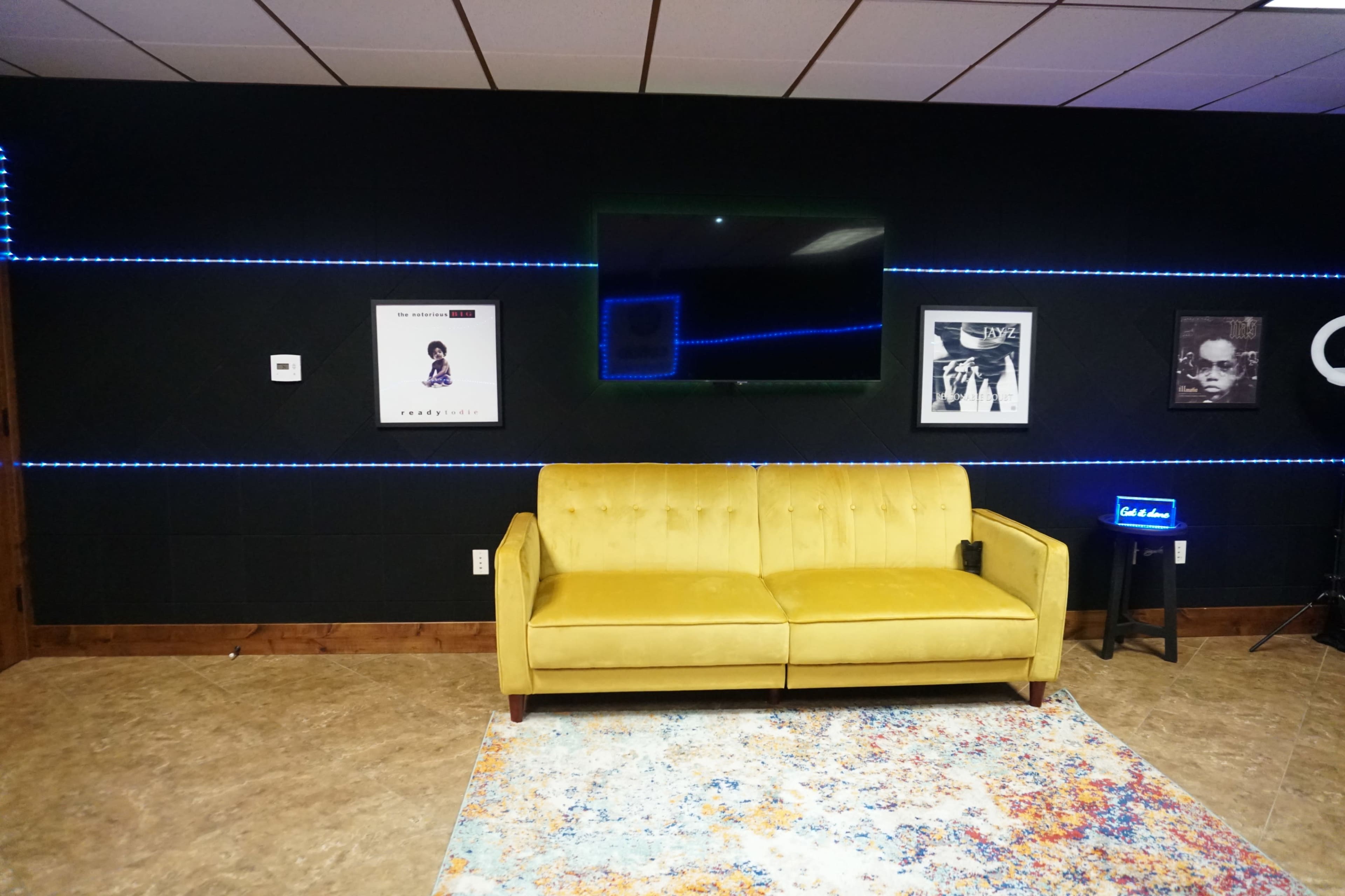 A yellow sofa positioned against a black wall adorned with framed posters and illuminated by blue LED strip lights.