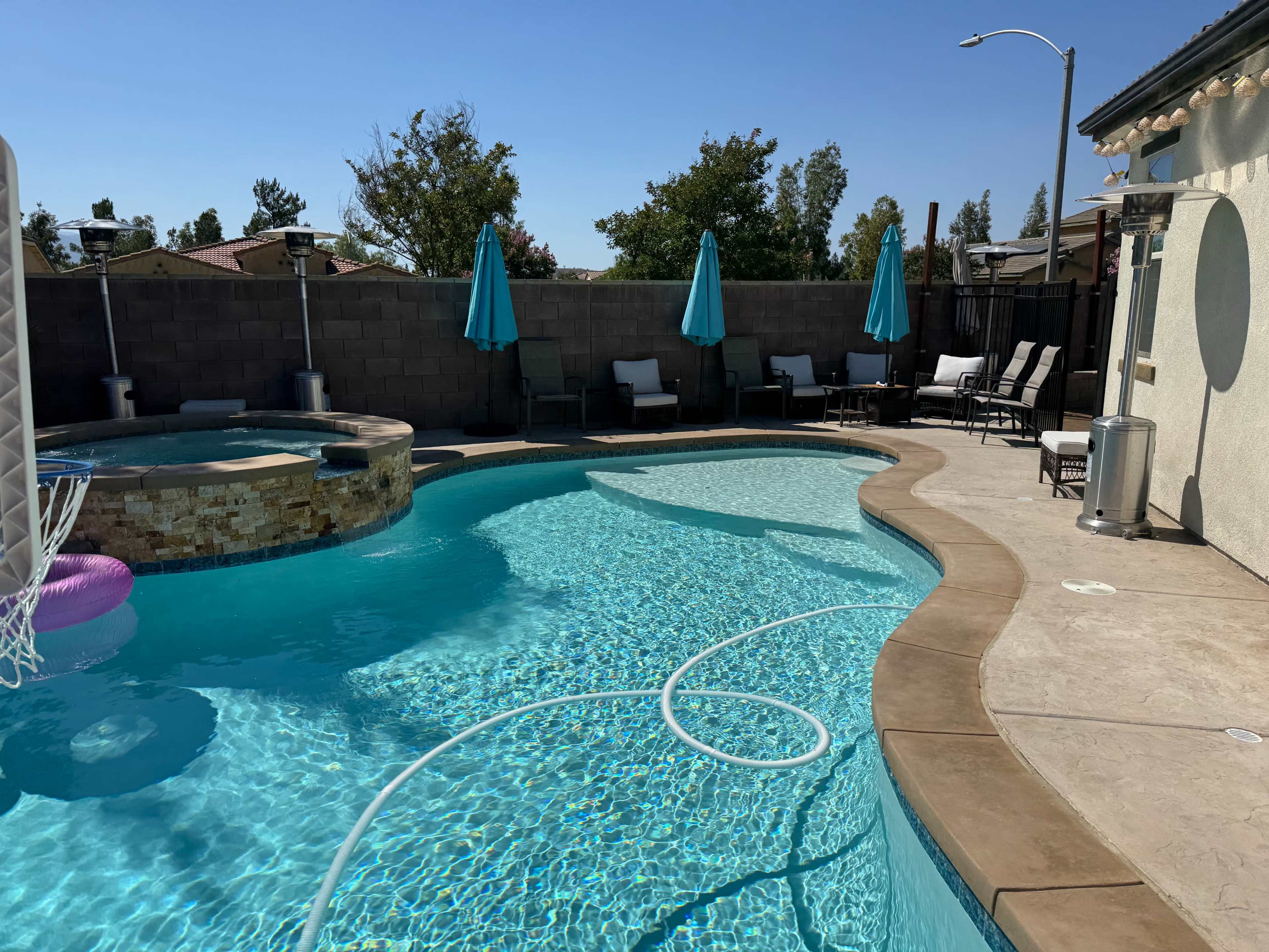 The image shows a swimming pool with a curved shape, surrounded by lounge chairs and umbrellas, along with a spa area, under a clear blue sky.