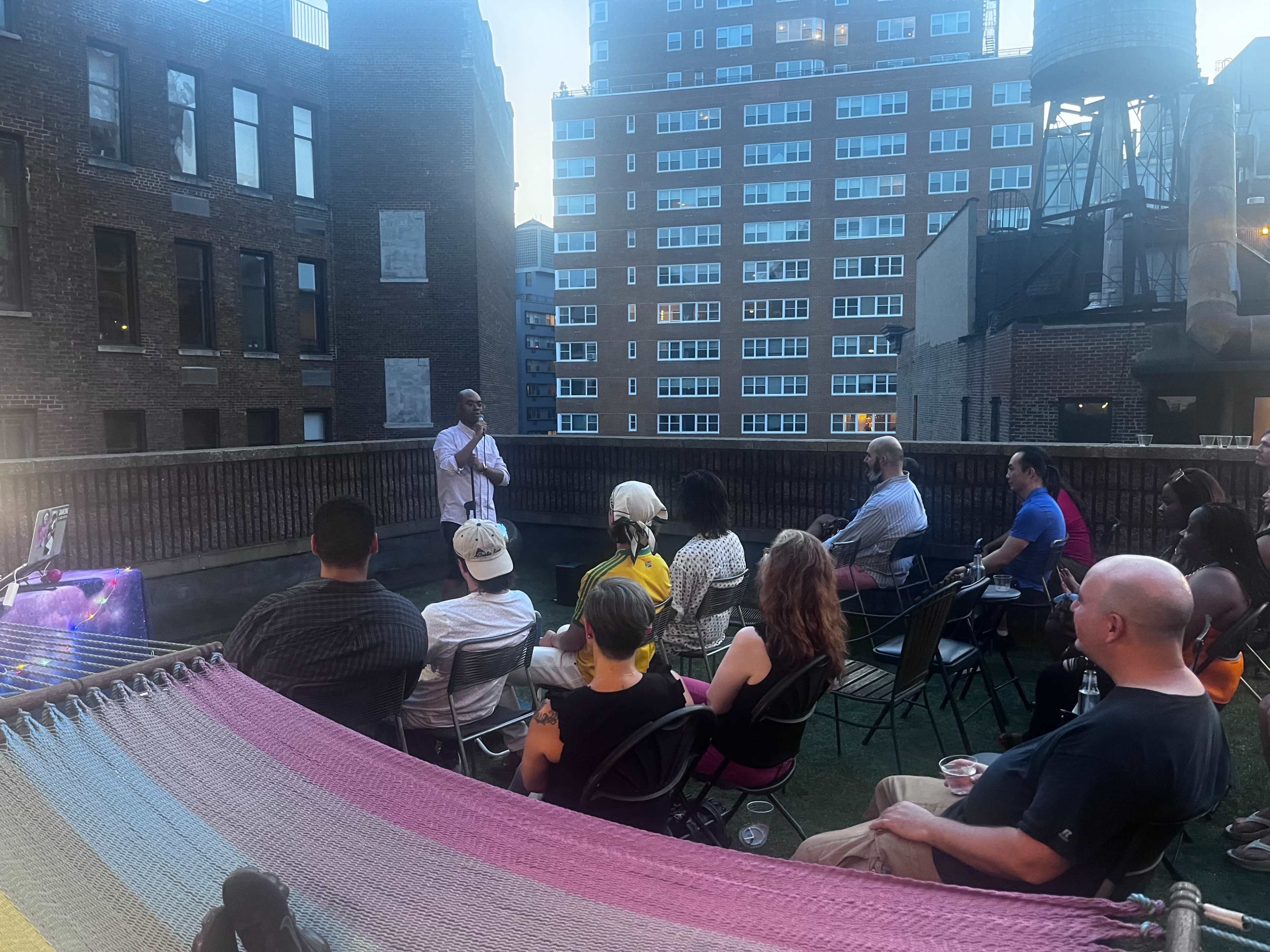 A group of people is seated in chairs on a rooftop, facing a speaker who is presenting near a hammock.
