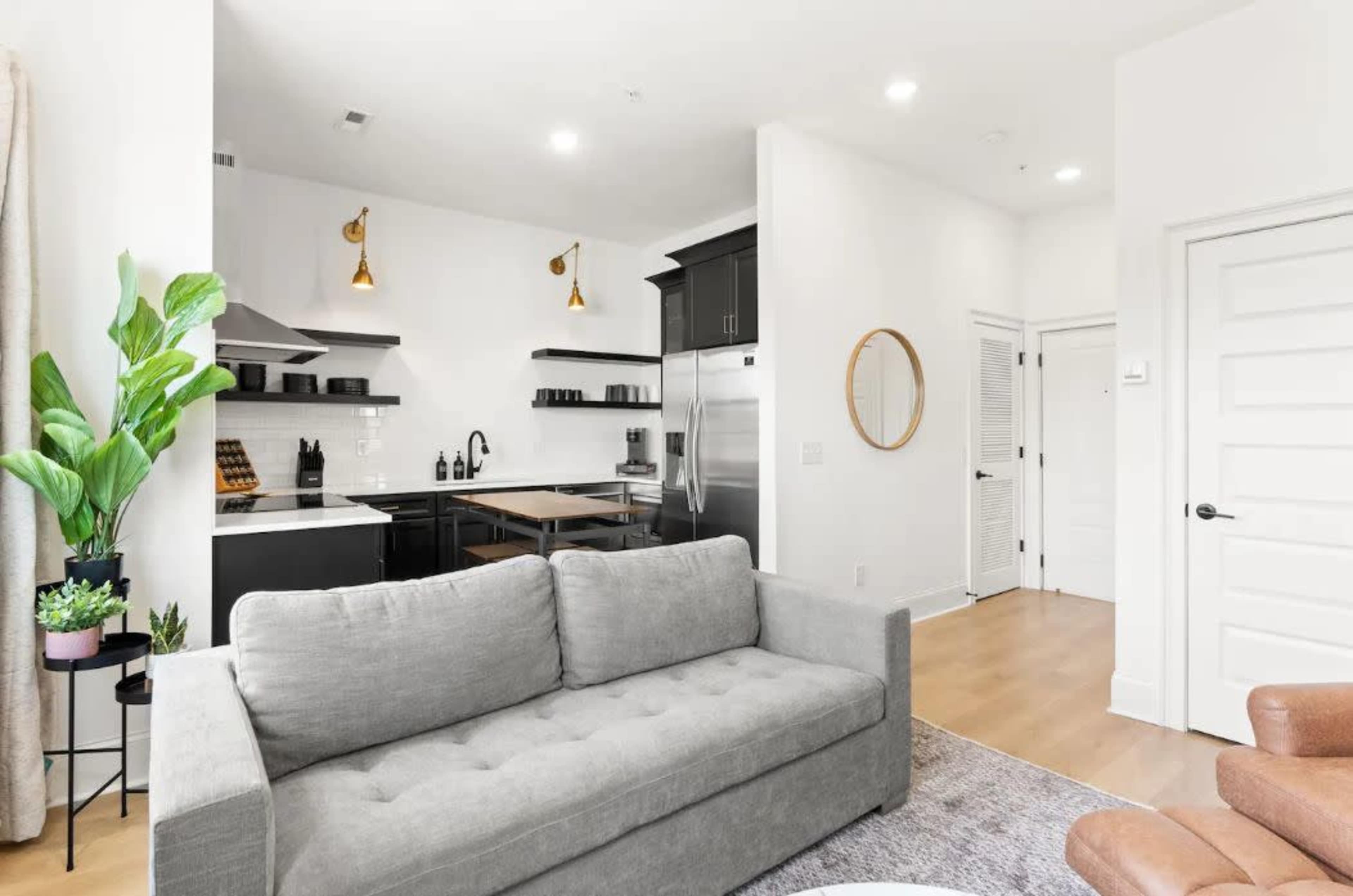 The image shows a modern living area with a gray sofa, a kitchen featuring stainless steel appliances, and a large indoor plant in a corner.