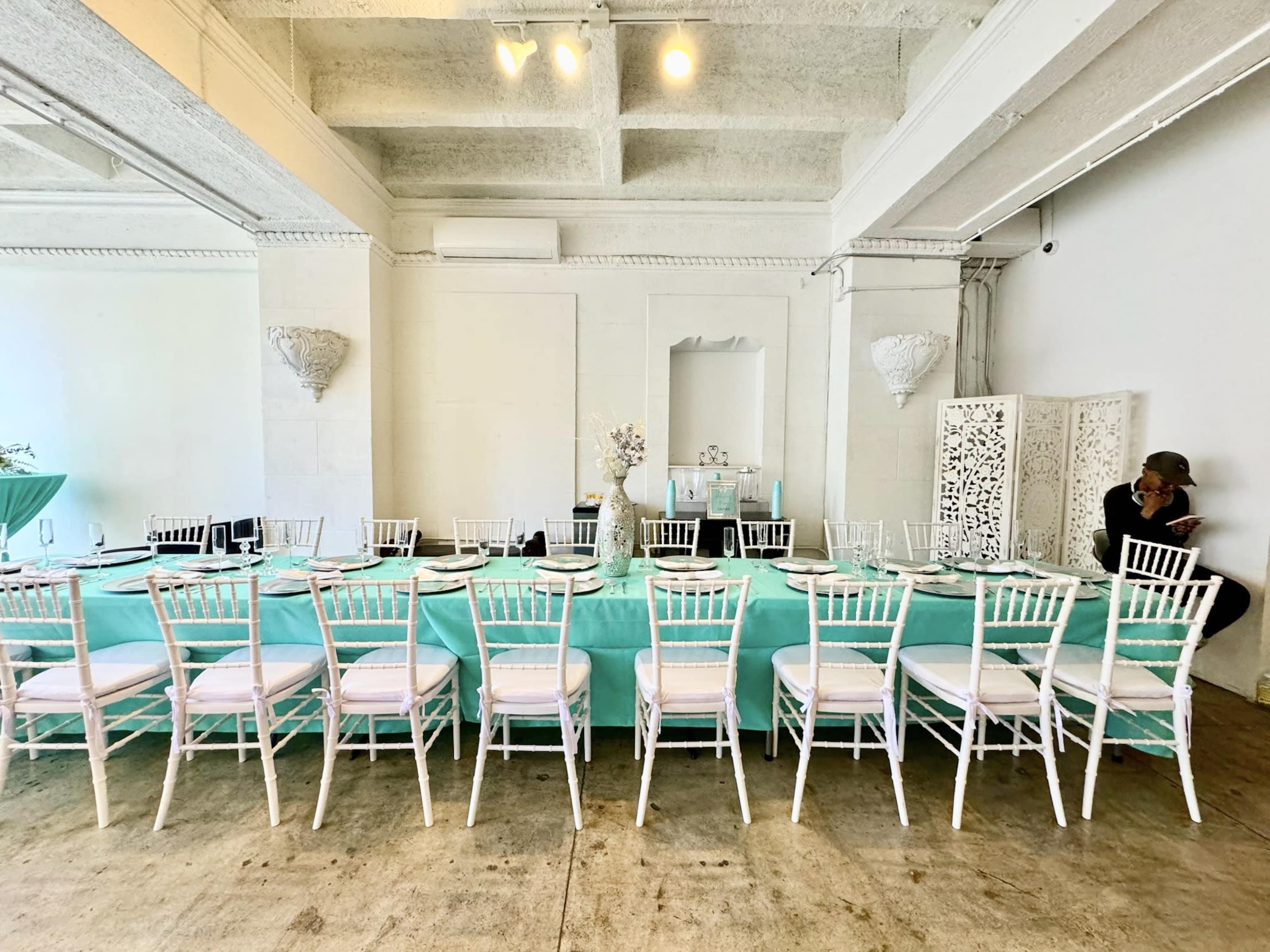 A long dining table with a turquoise tablecloth is set with white chairs in a bright, minimalistic room.
