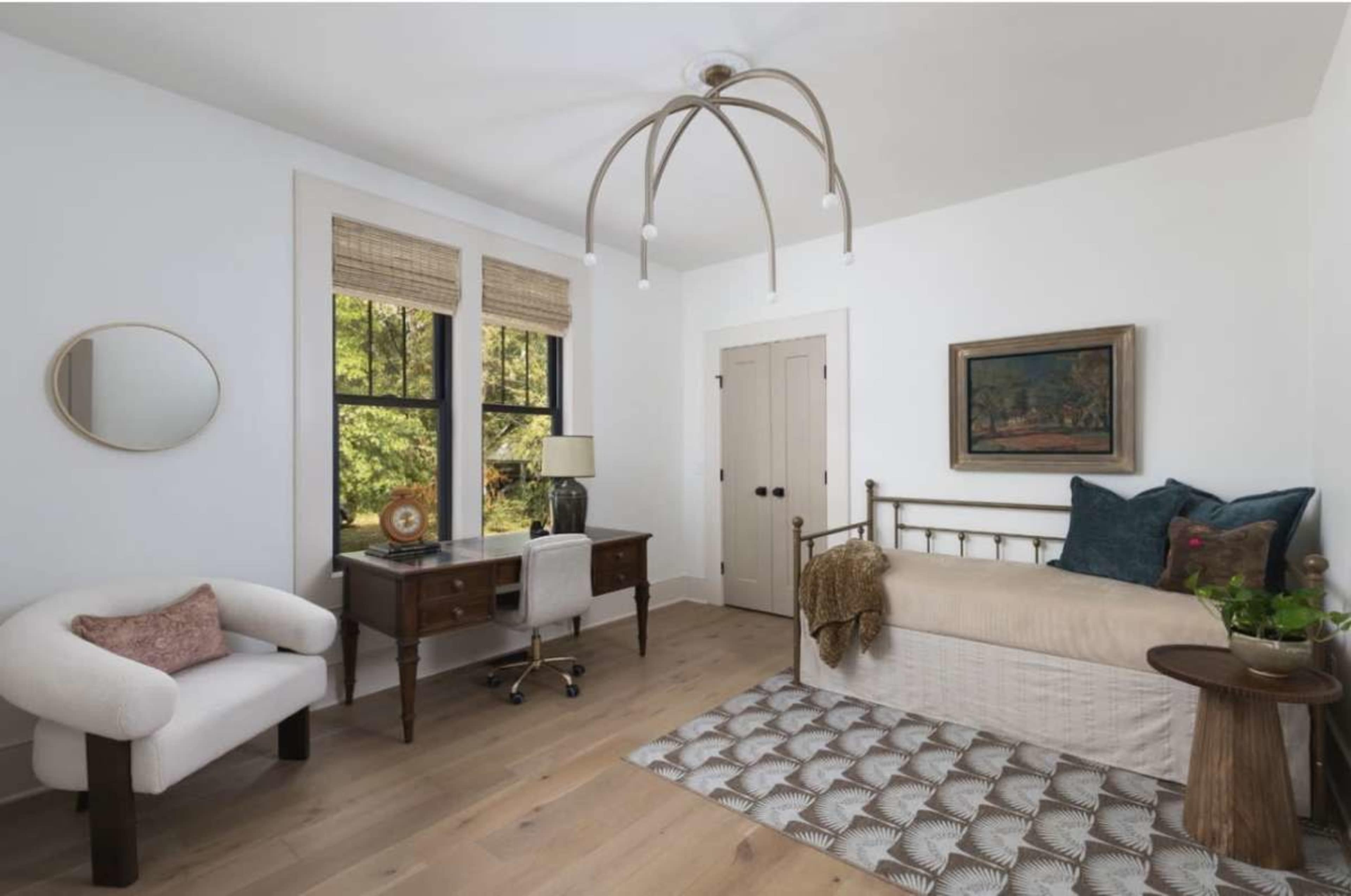 The image shows a well-lit room featuring a bed, a desk with a chair, and a small seating area, all arranged around a patterned rug.