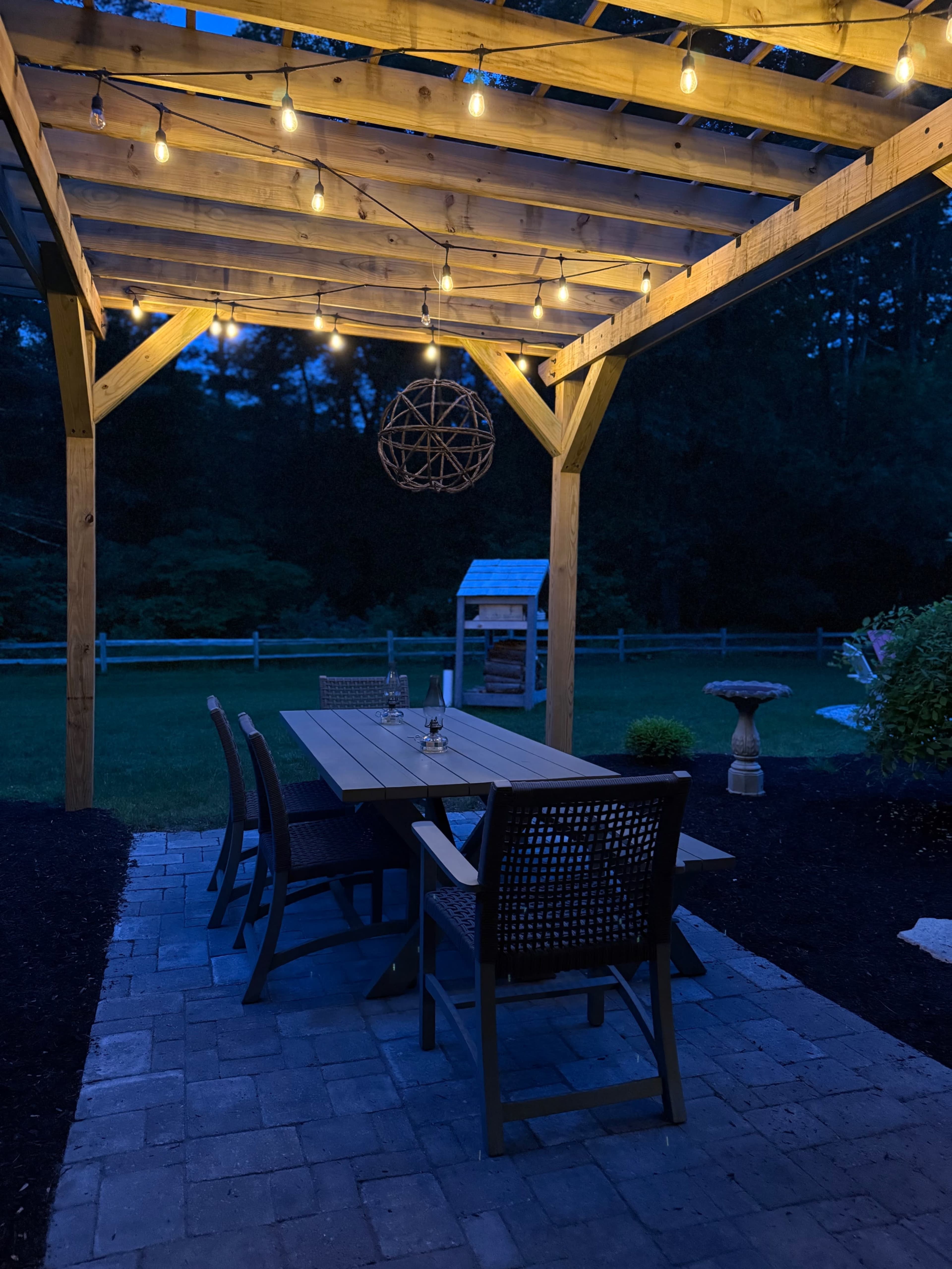 A wooden pergola with string lights illuminates a patio table and chairs in a backyard setting during dusk.