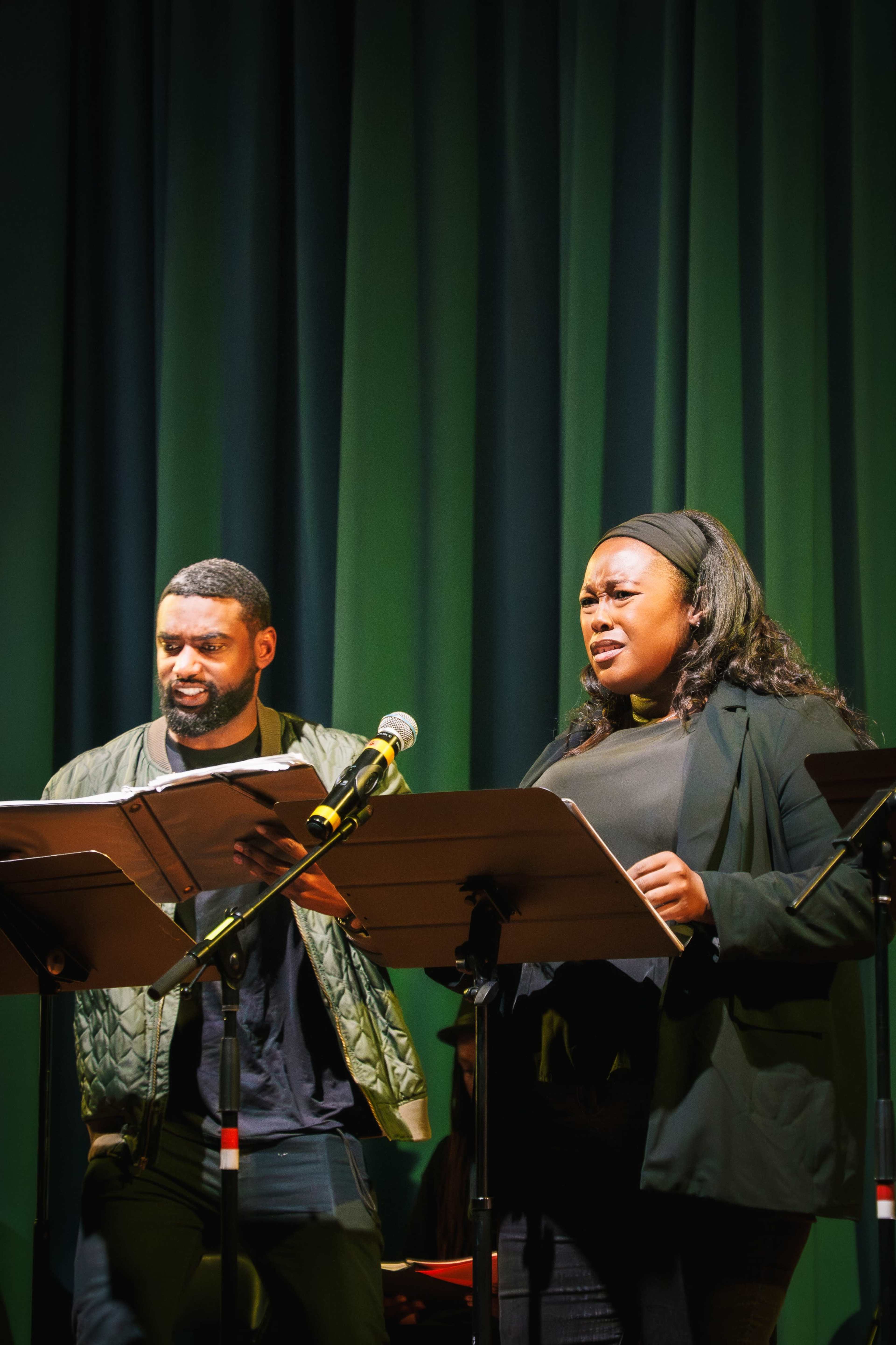 Two performers stand on stage, reading from scripts into microphones against a green curtain backdrop.