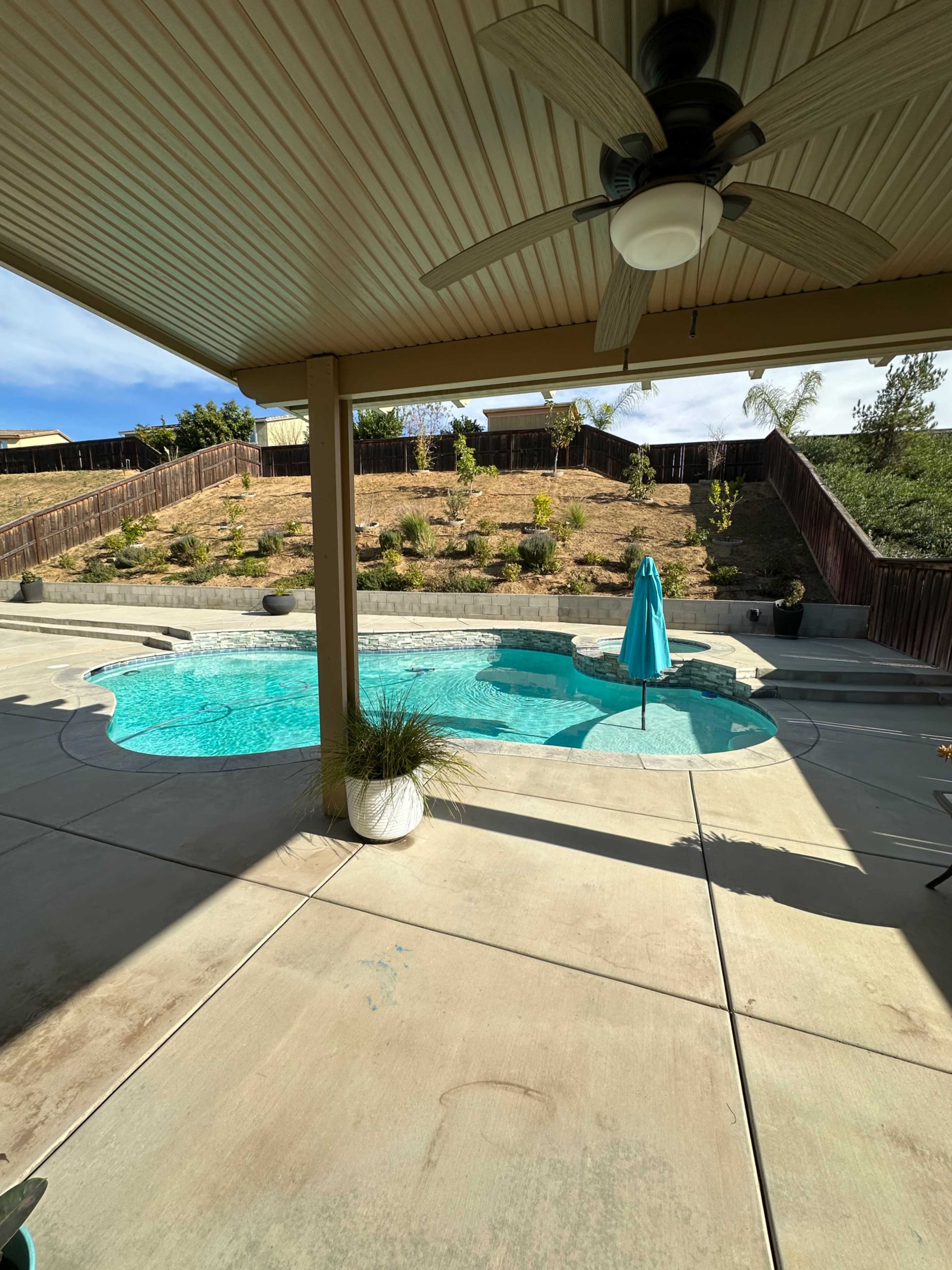 The image shows a swimming pool area with a turquoise umbrella, surrounded by a landscaped yard and a concrete patio under a shaded roof.