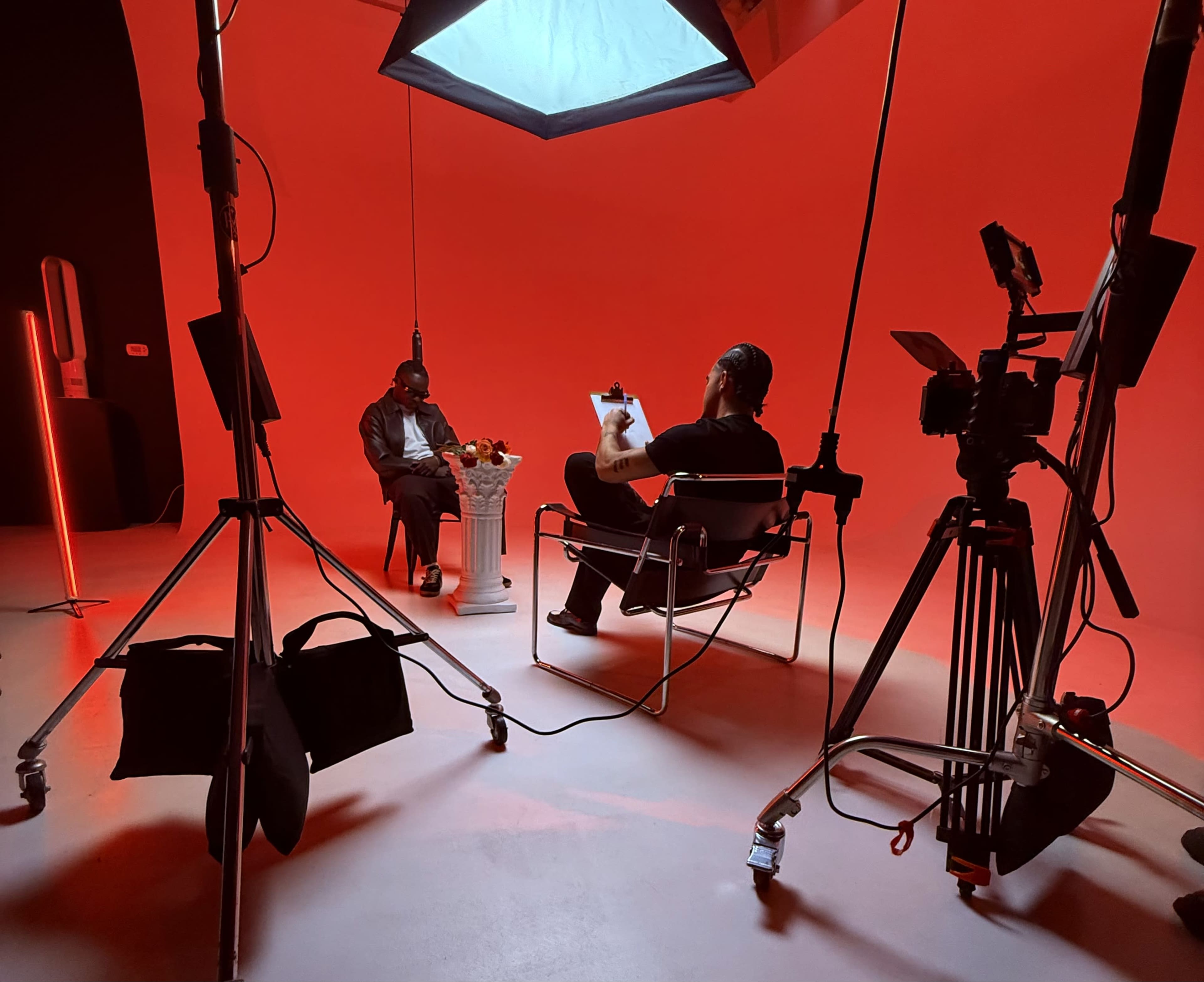A seated interview is taking place in a studio with a red backdrop, featuring two people, one in a chair sketching and the other engaged in conversation.