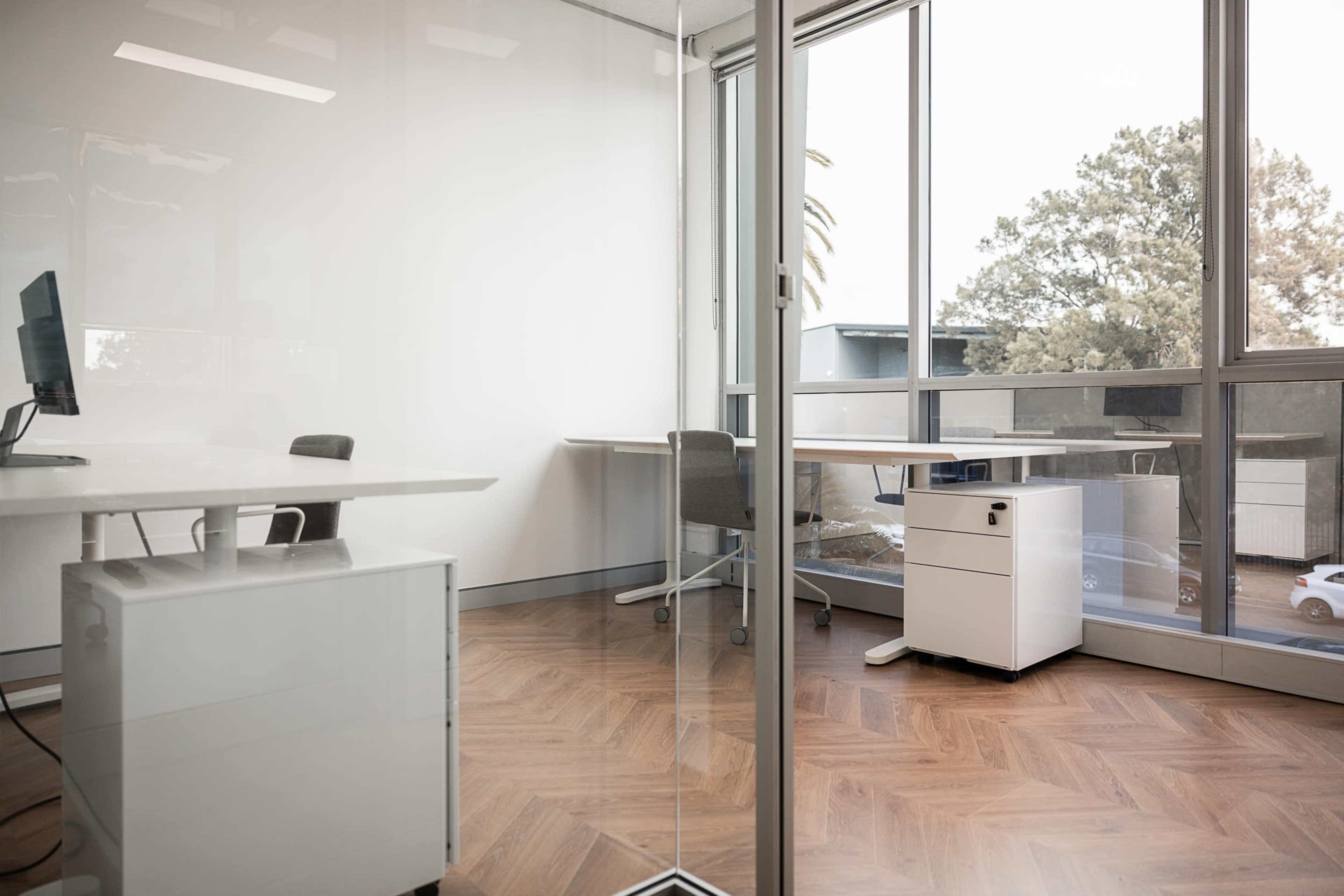 A modern office space with a glass partition, featuring a desk with a computer on one side and a white filing cabinet on the other.