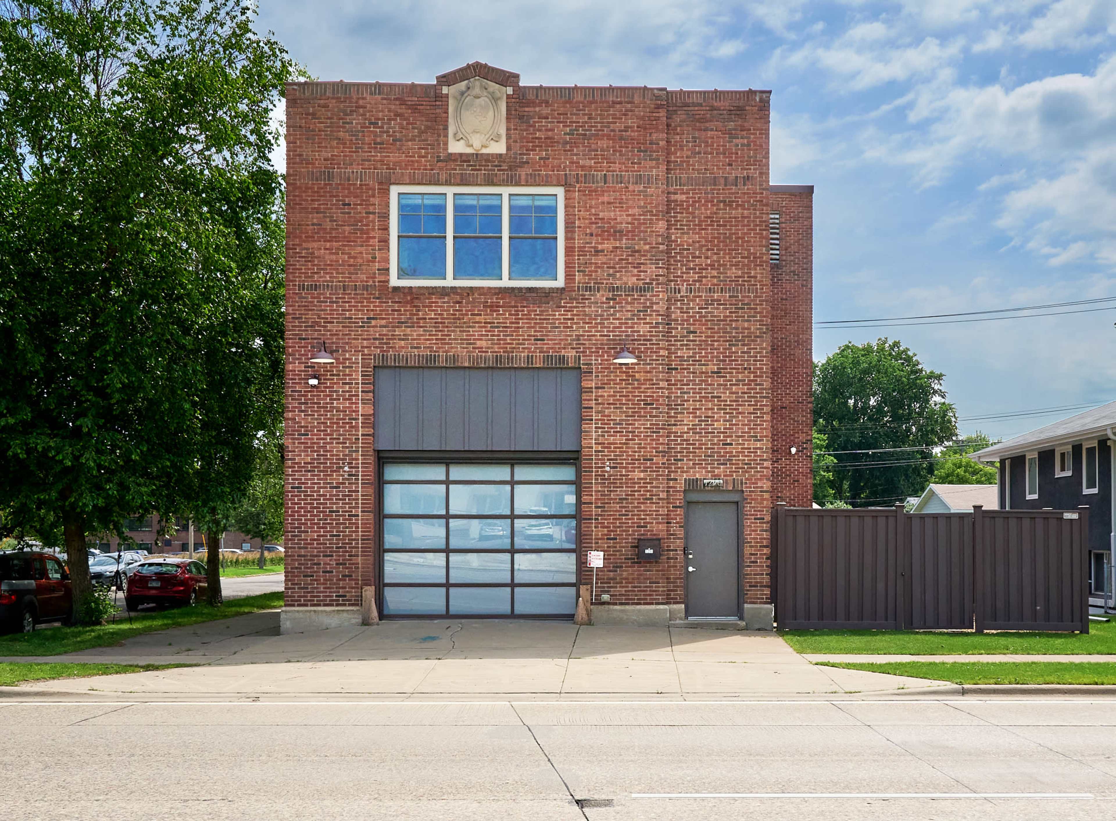 The image shows a two-story brick building with large windows and a garage door, situated on a paved street with green grass and a fence alongside it.