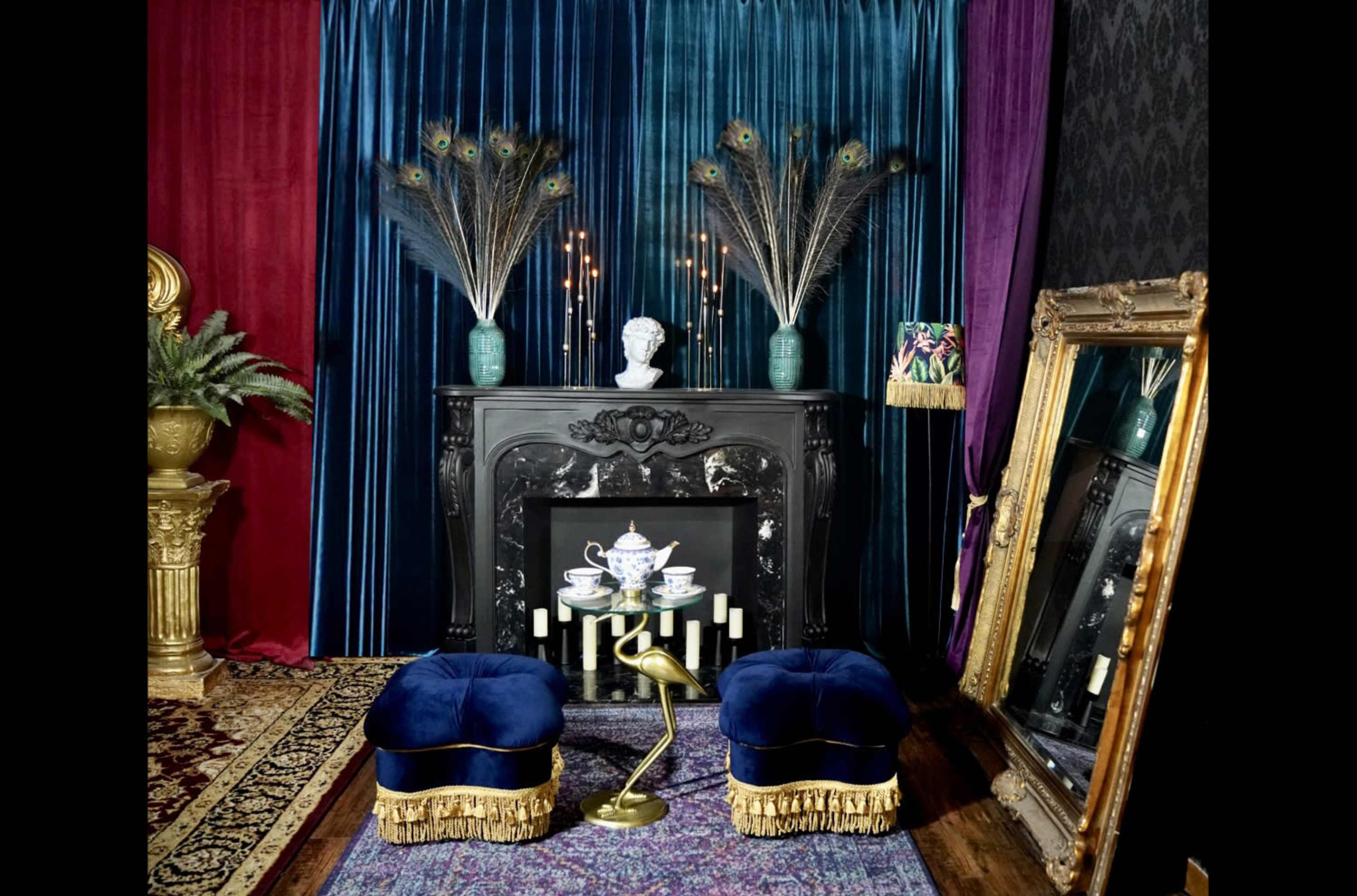 The scene features a richly decorated interior with a dark fireplace adorned by a bust, peacock feathers in vases, and two blue velvet stools in front of an ornate mirror.