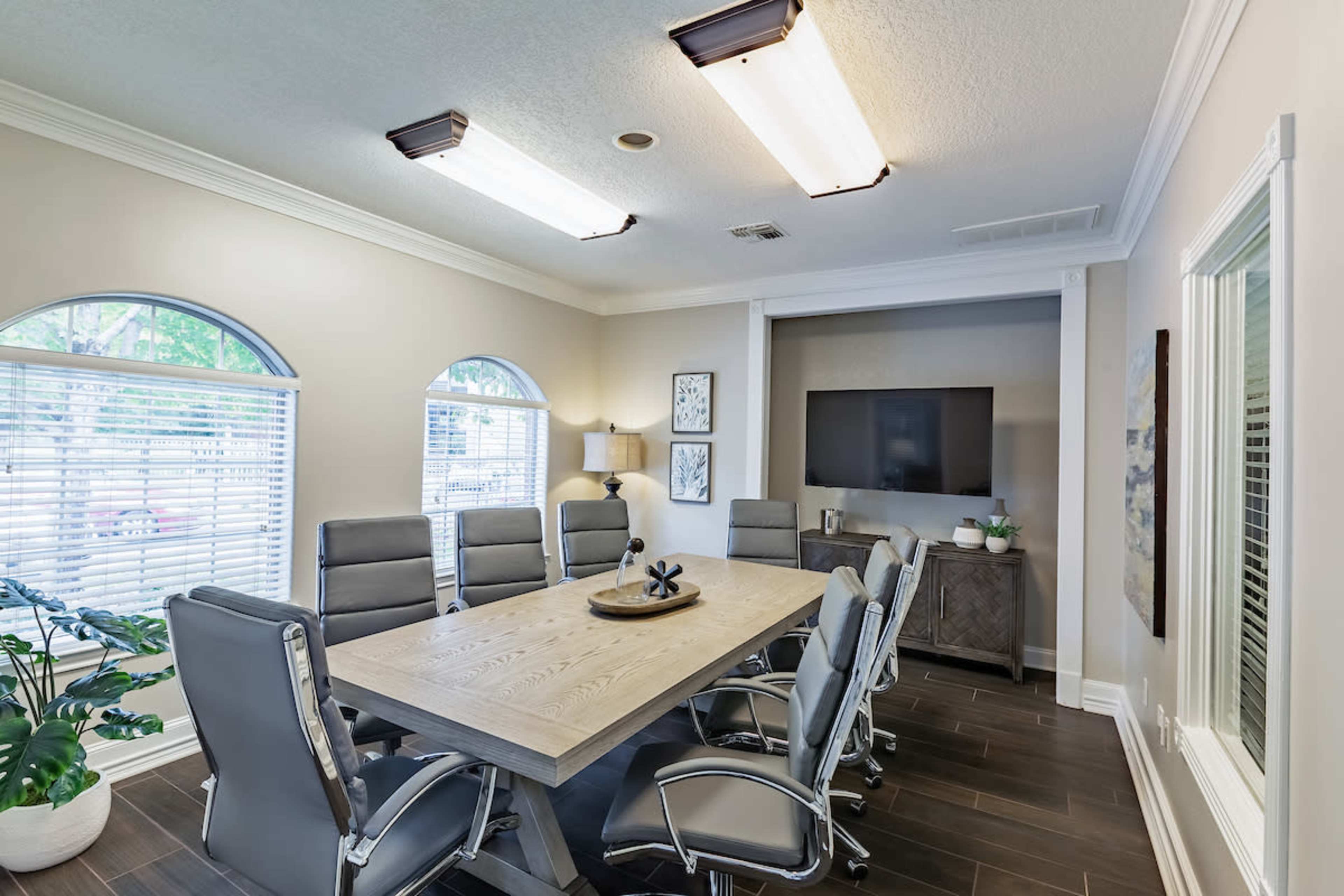 A conference room features a large wooden table surrounded by six gray chairs, with a television on the wall and natural light coming through arched windows.