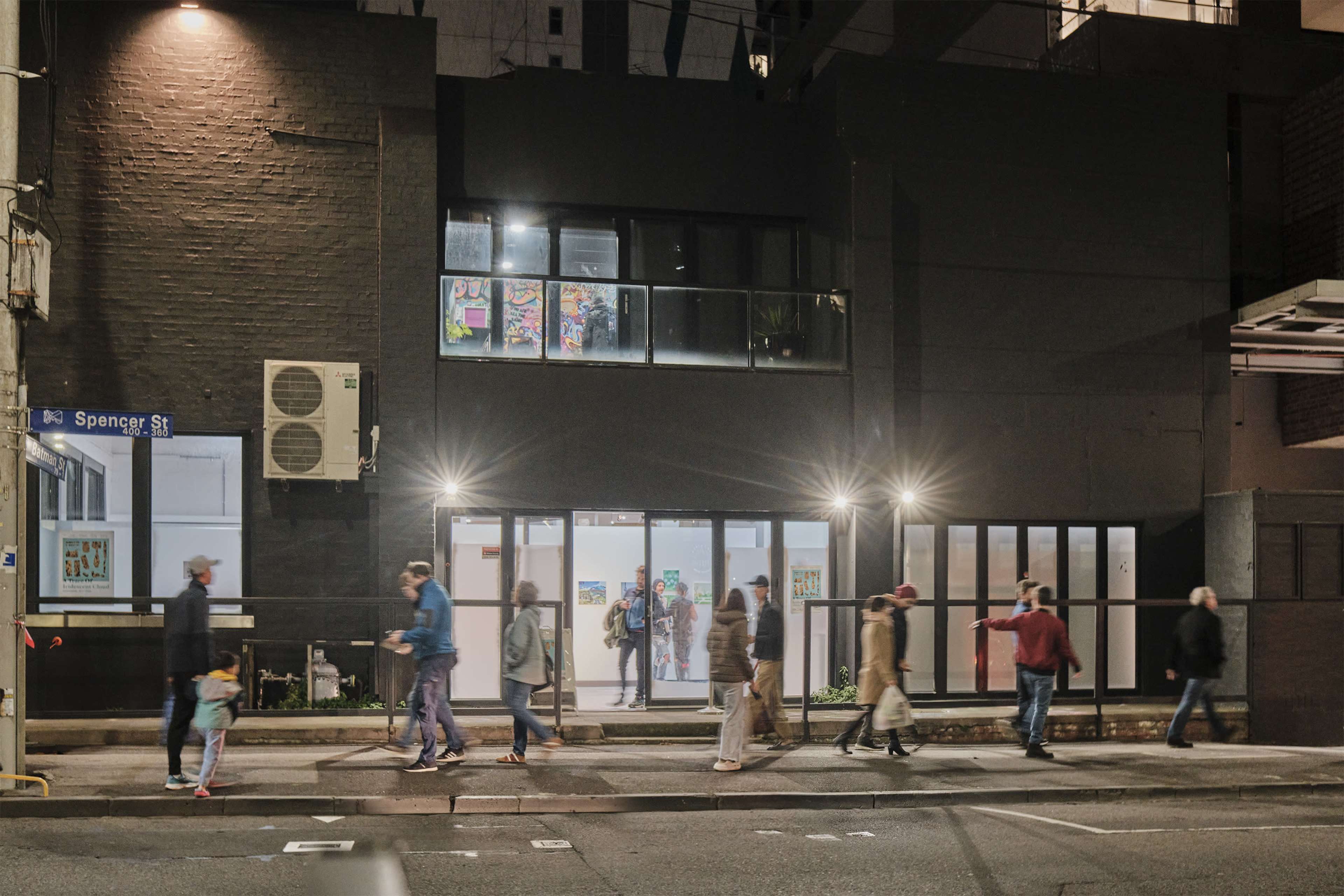 A group of people walks along a sidewalk in front of a modern black building with illuminated windows at night.