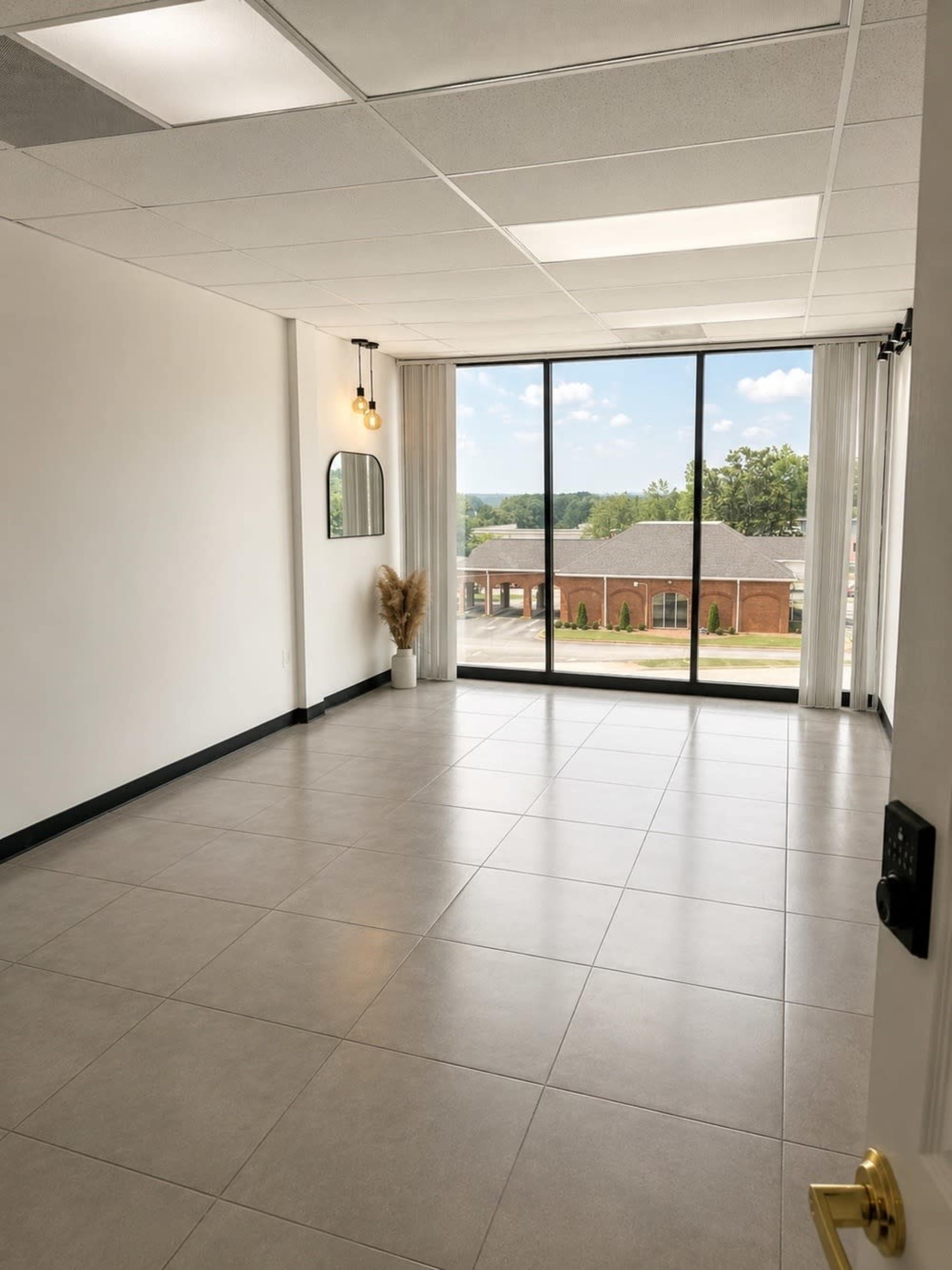 The image shows an empty room with large windows overlooking a street, featuring gray tile flooring and minimal decor.
