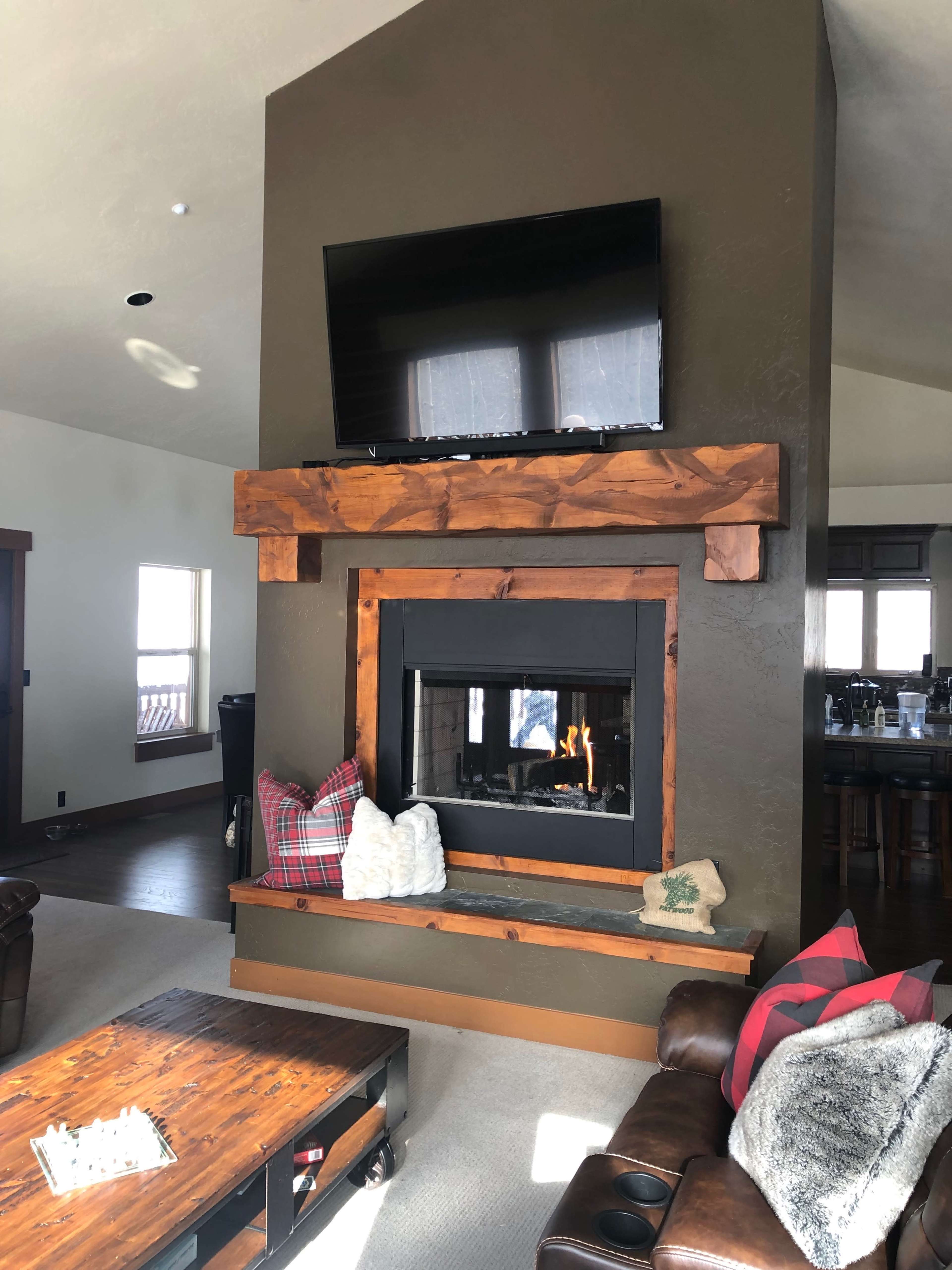 A living room with a large wooden mantel, a flat-screen TV mounted above a fireplace, and a comfortable seating area with pillows and a coffee table.