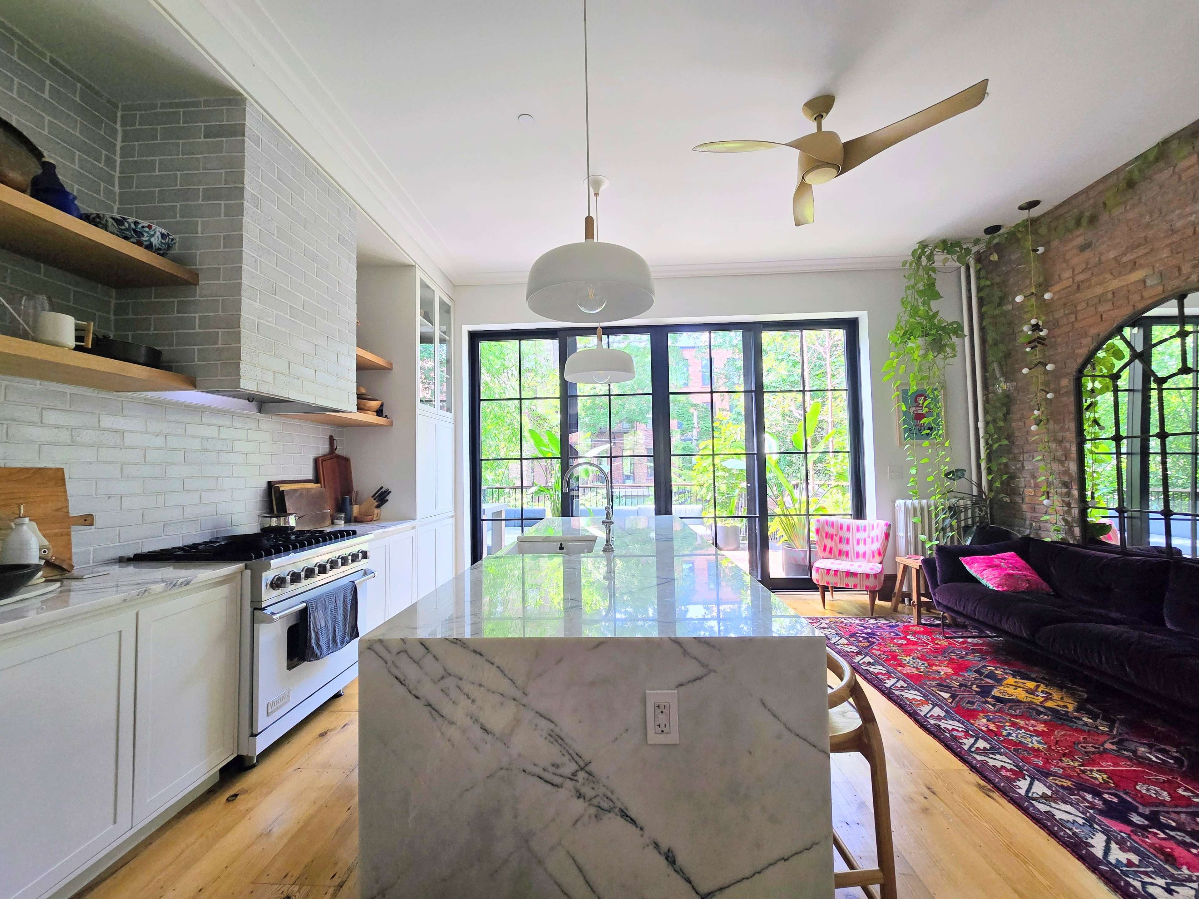 The kitchen features a large marble island, modern appliances, and large windows overlooking a green outdoor space.