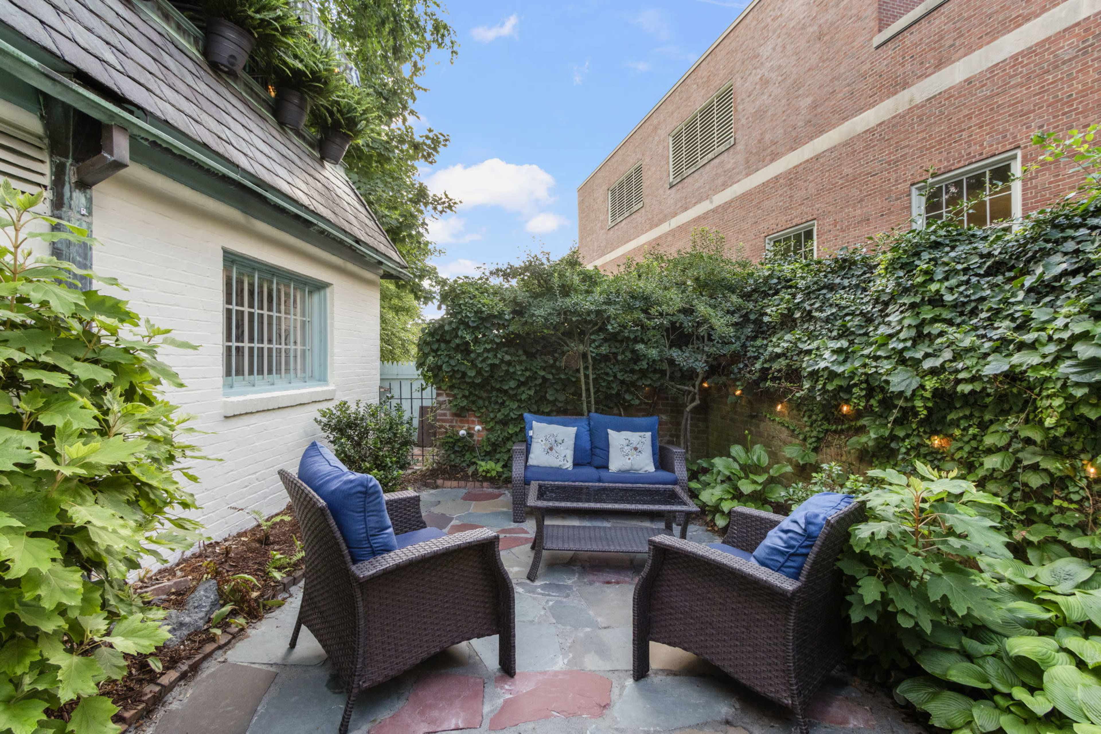 The image shows a small outdoor patio area with stone flooring, featuring a seating arrangement of two chairs and a sofa surrounded by greenery and ivy-covered walls.