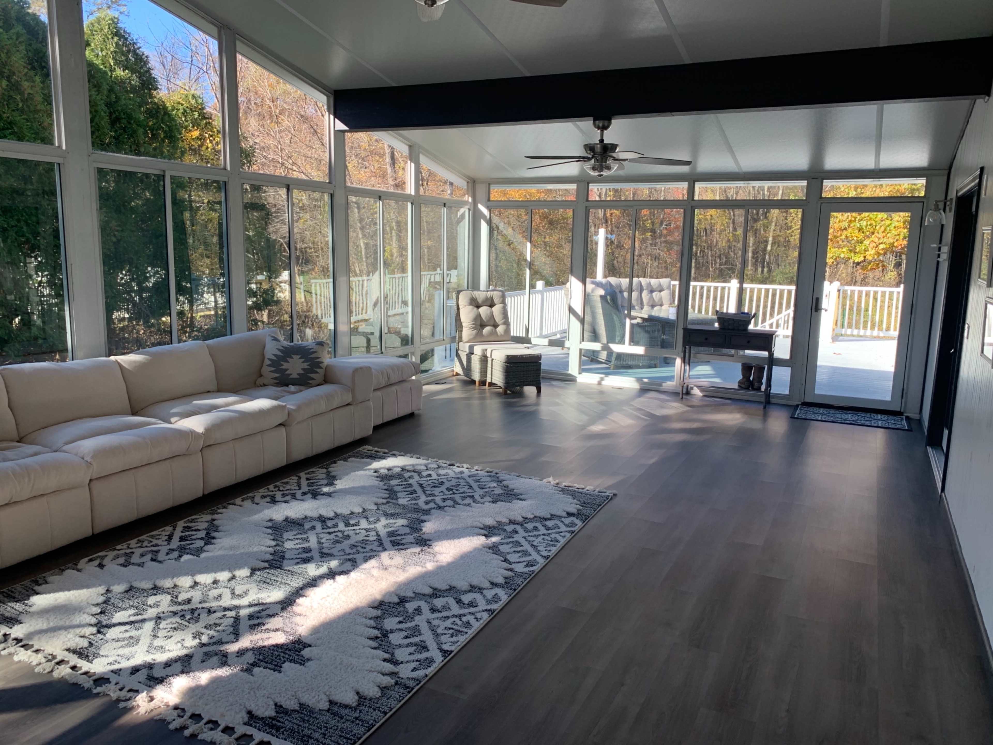 A sunlit sunroom featuring a beige sectional sofa, a patterned area rug, and large windows overlooking a wooded area.