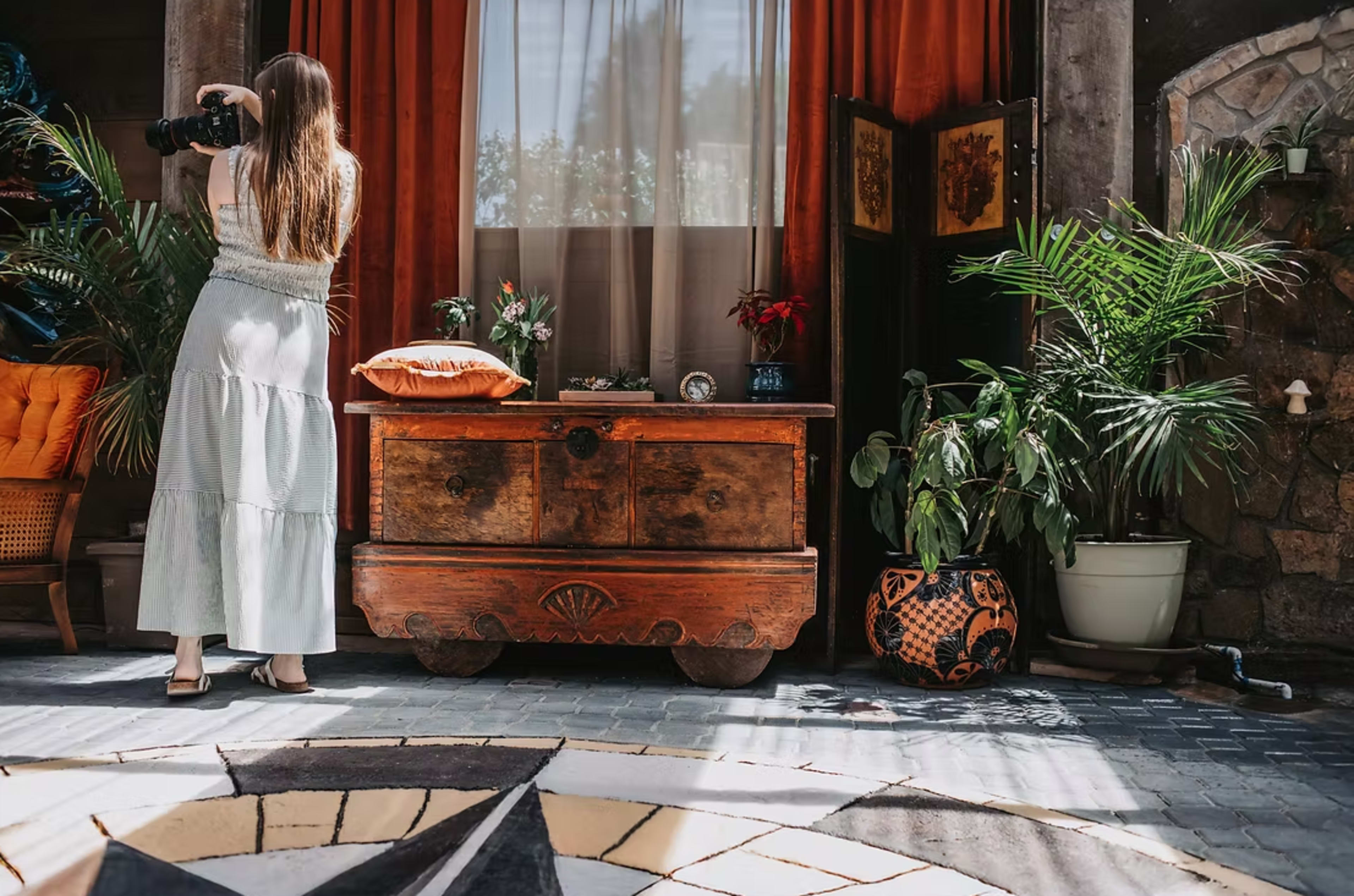 A woman is photographing a space with an antique wooden chest amid plants and vintage decor.
