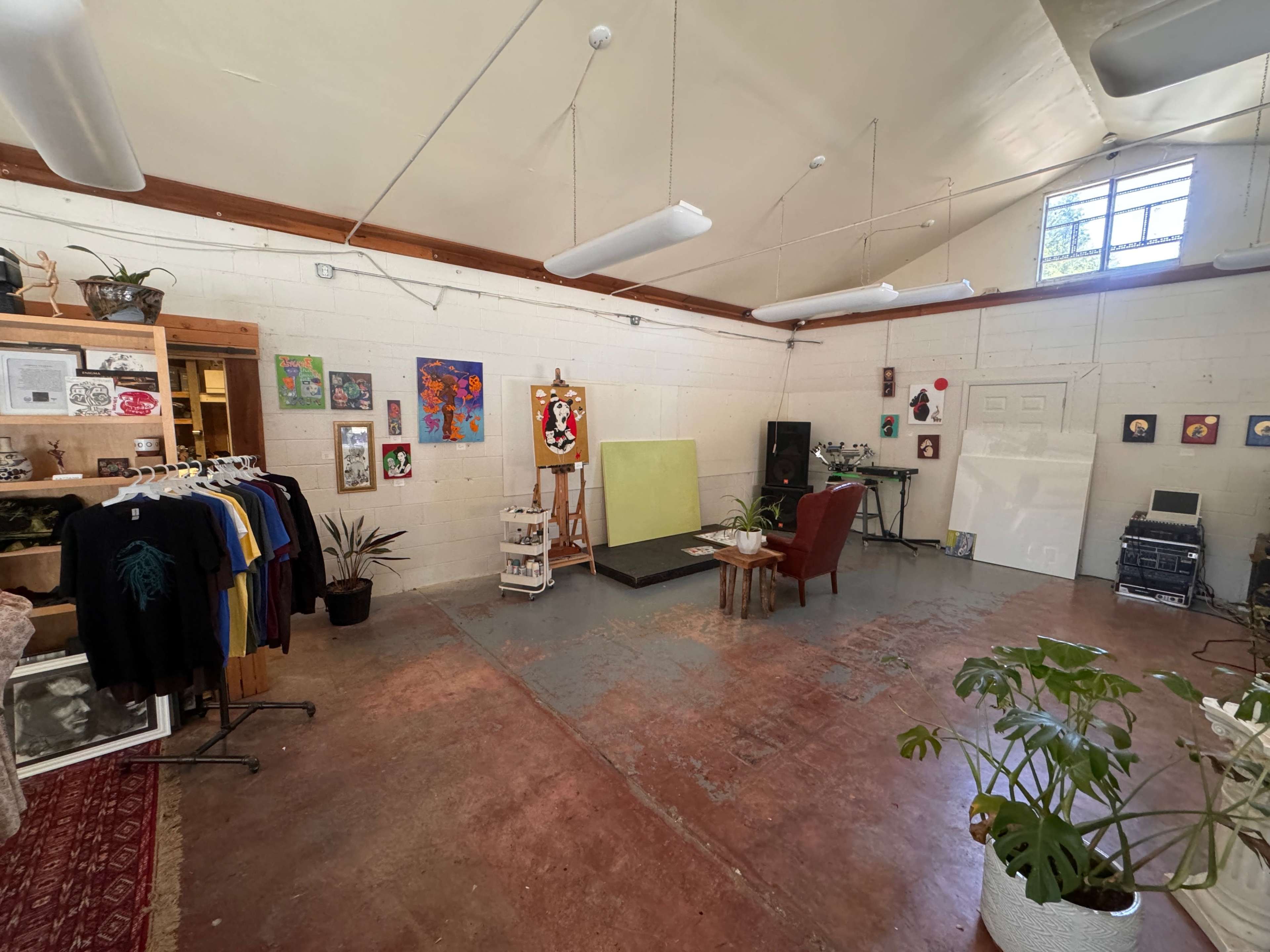 An art studio featuring clothing racks, several paintings on the walls, an easel with a blank canvas, a chair, and various plants.