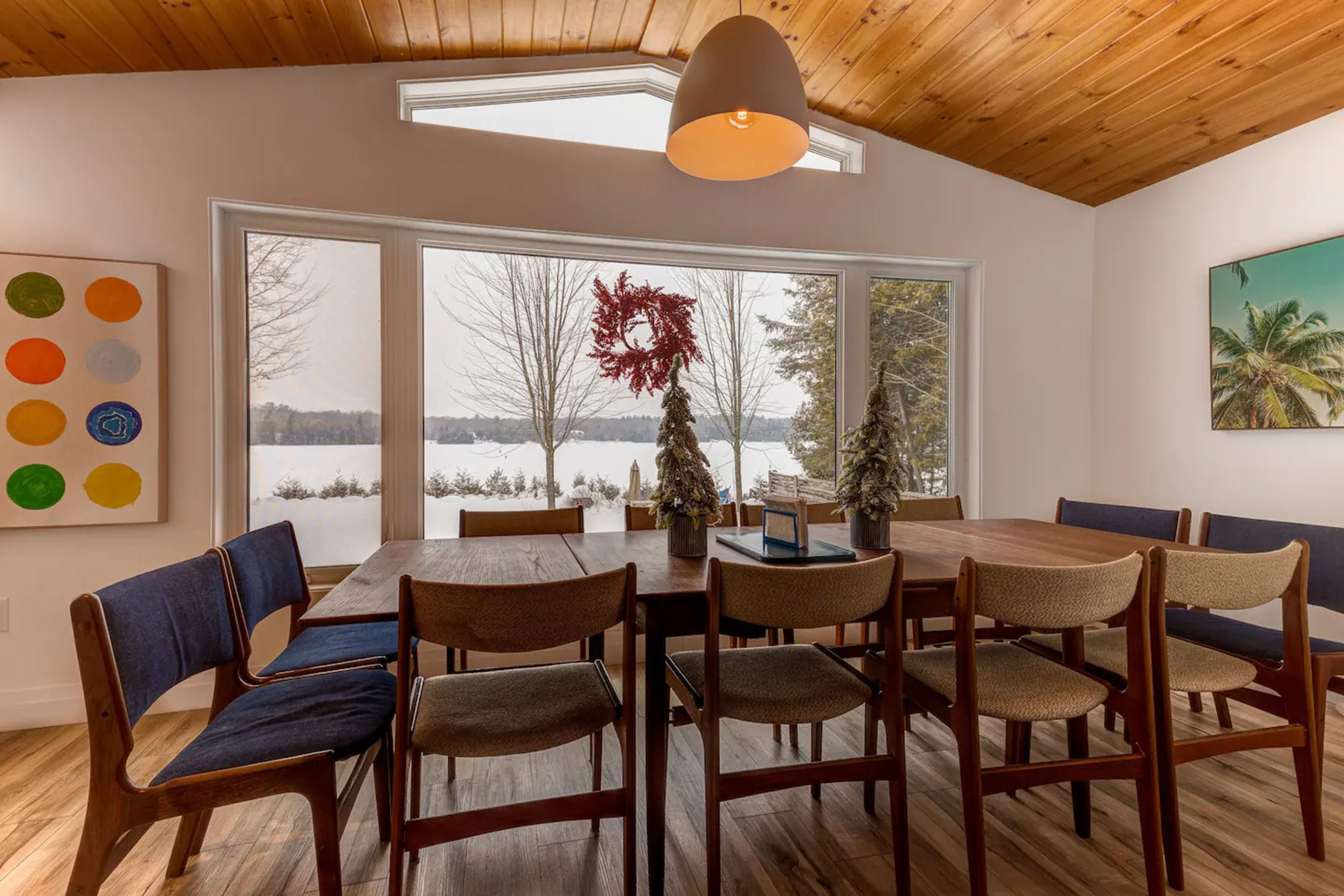A dining area features a wooden table surrounded by chairs, with large windows offering a view of a snowy landscape outside.