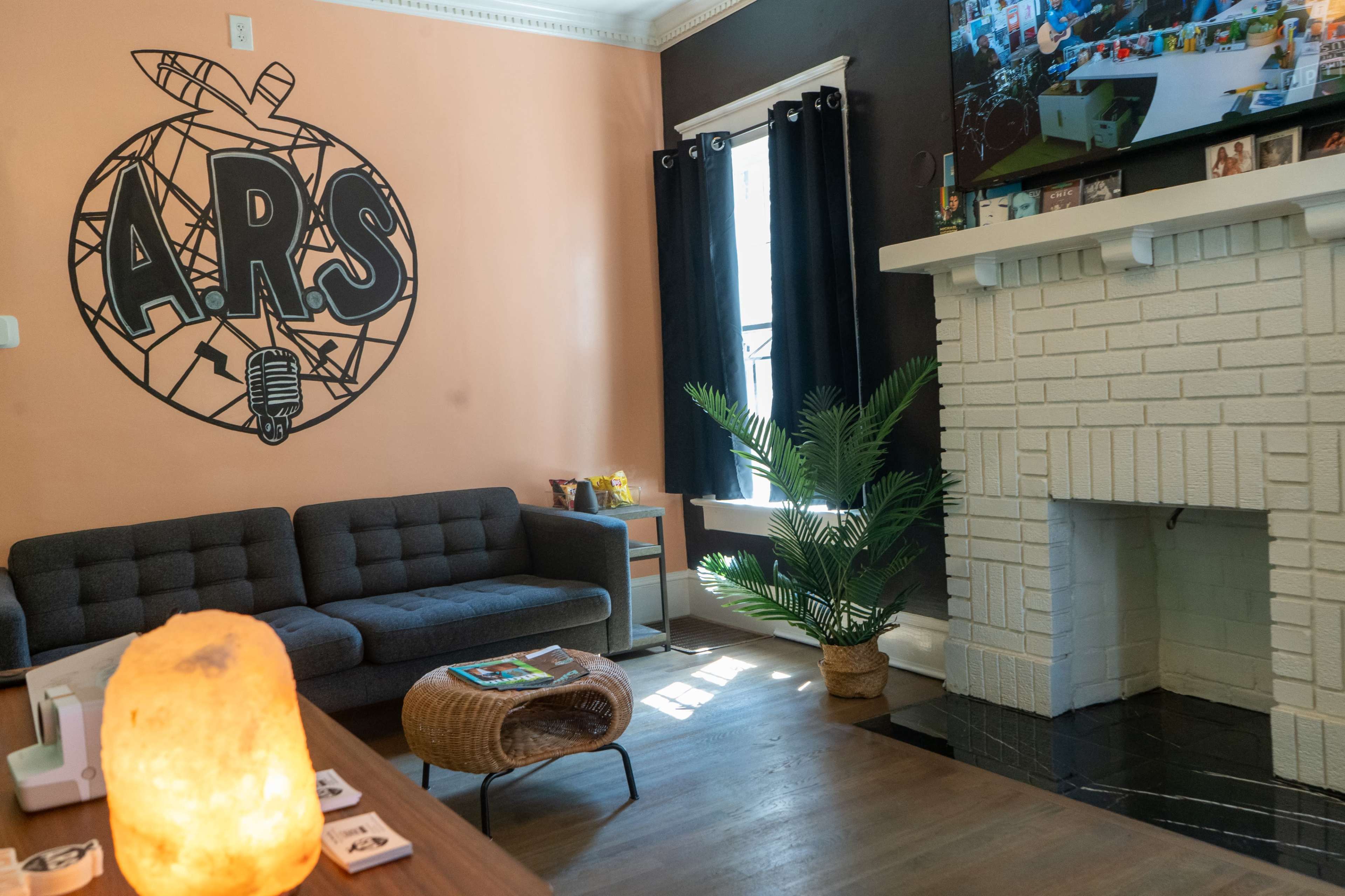 The image shows a cozy lounge area featuring a gray sofa, a decorative wall with the letters "A.R.S.," a plant beside a white fireplace, and a wooden coffee table with magazines.