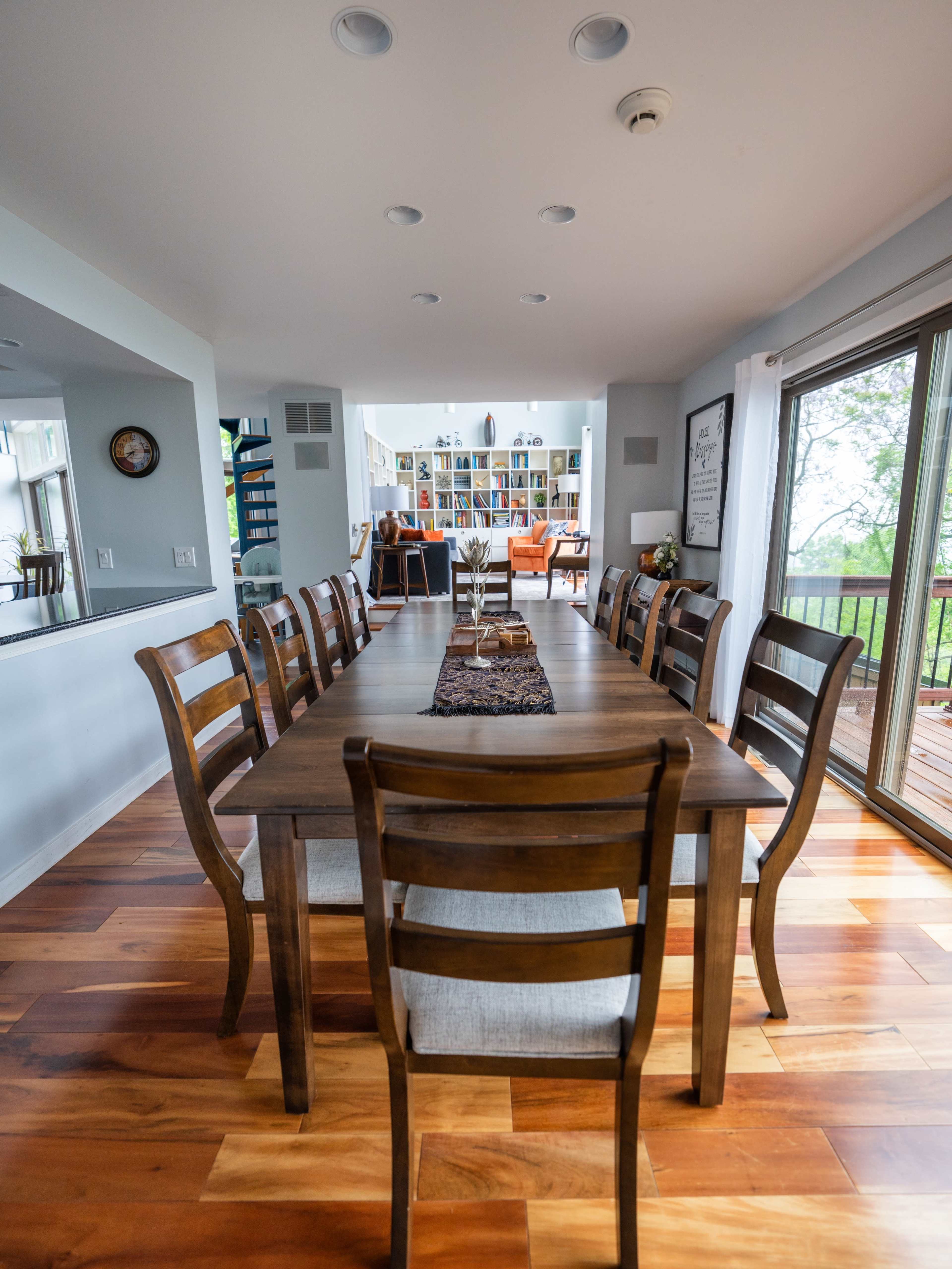A long wooden dining table surrounded by chairs, with a decorative centerpiece, leading to a living area filled with bookshelves and large windows.