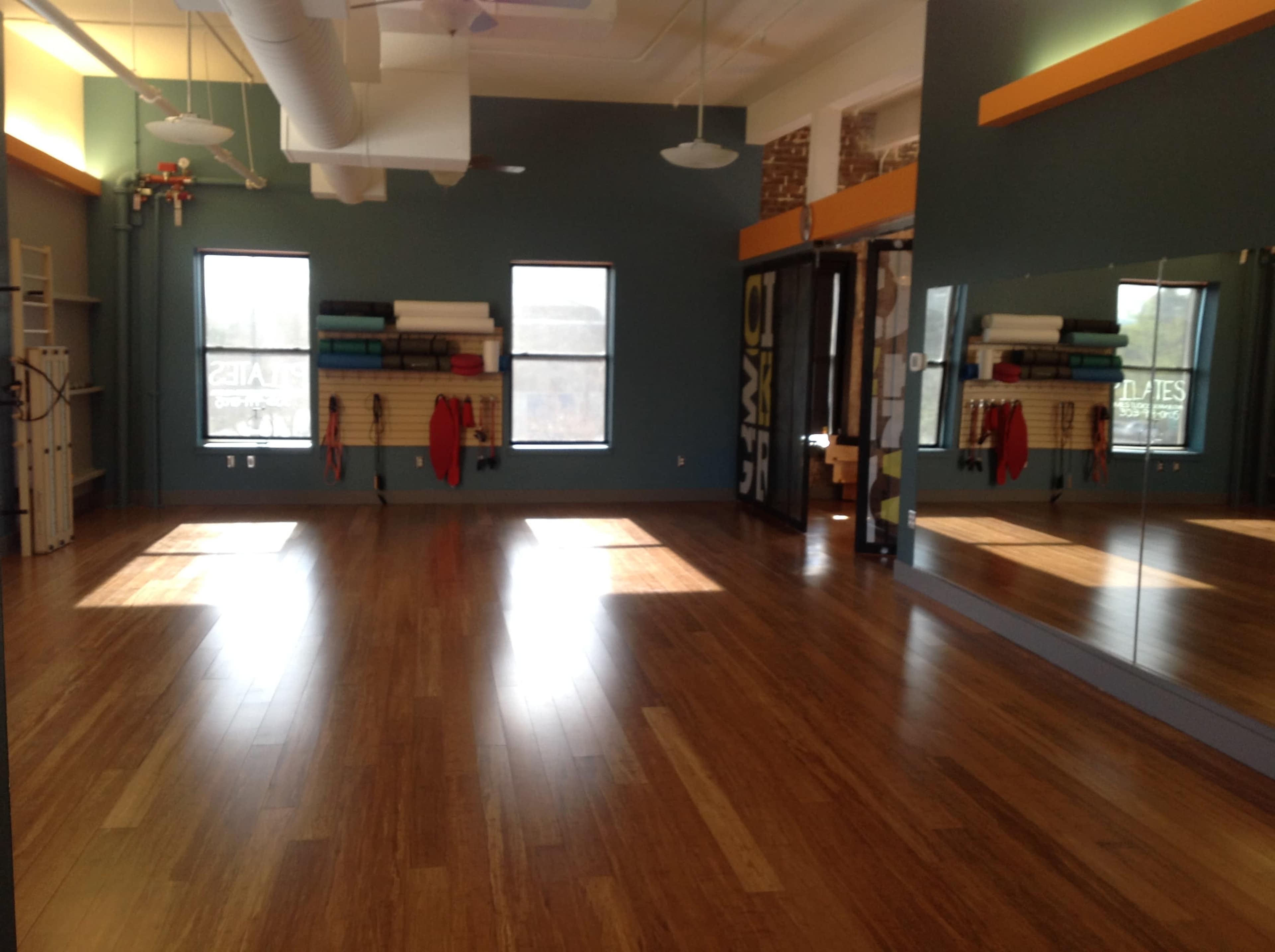 A spacious, empty studio with wooden flooring, large windows allowing natural light, and mirrors along one wall.