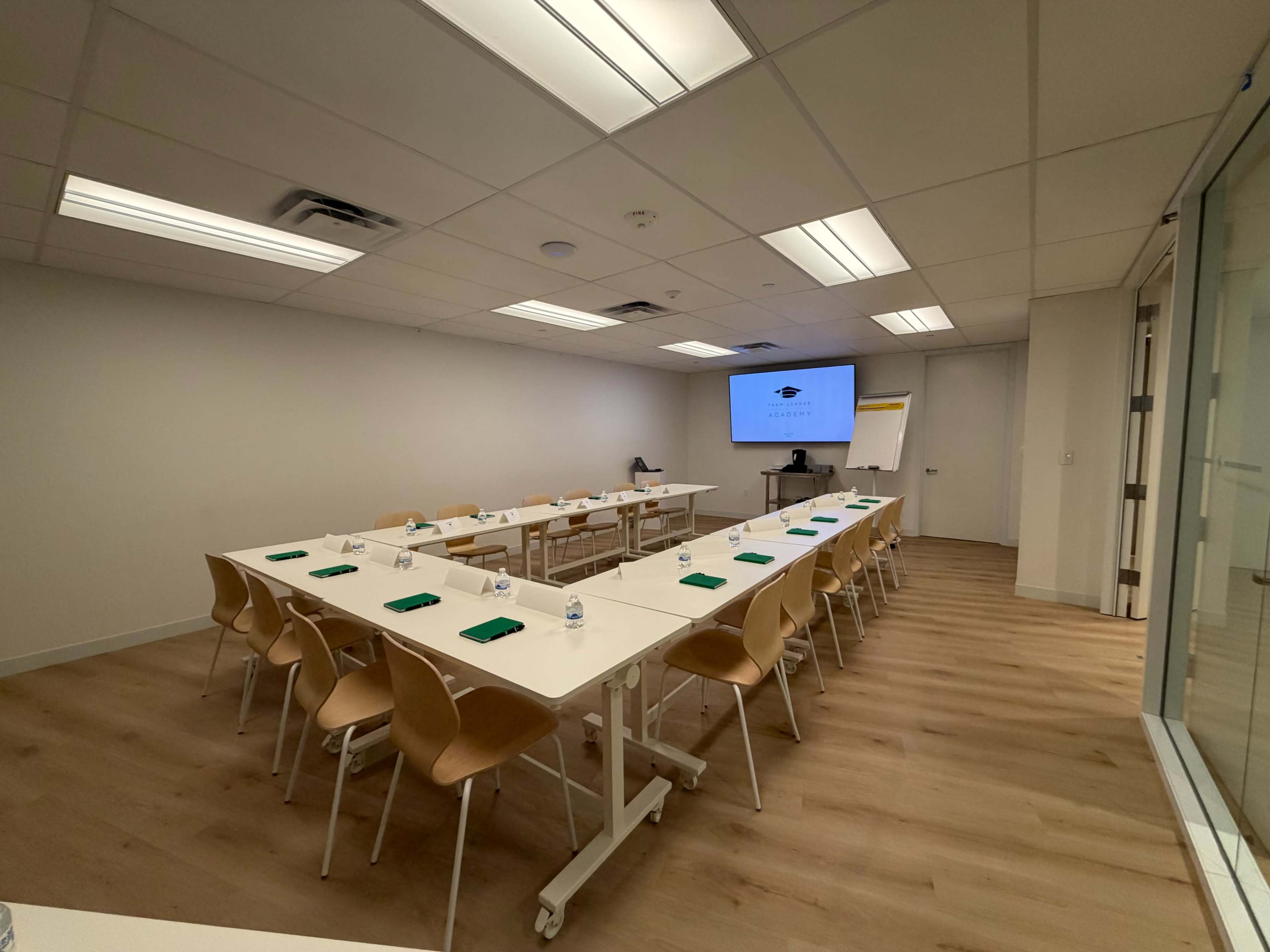 The image shows a modern conference room with a long table arranged with chairs, water bottles, and notepads, along with a presentation screen at one end.