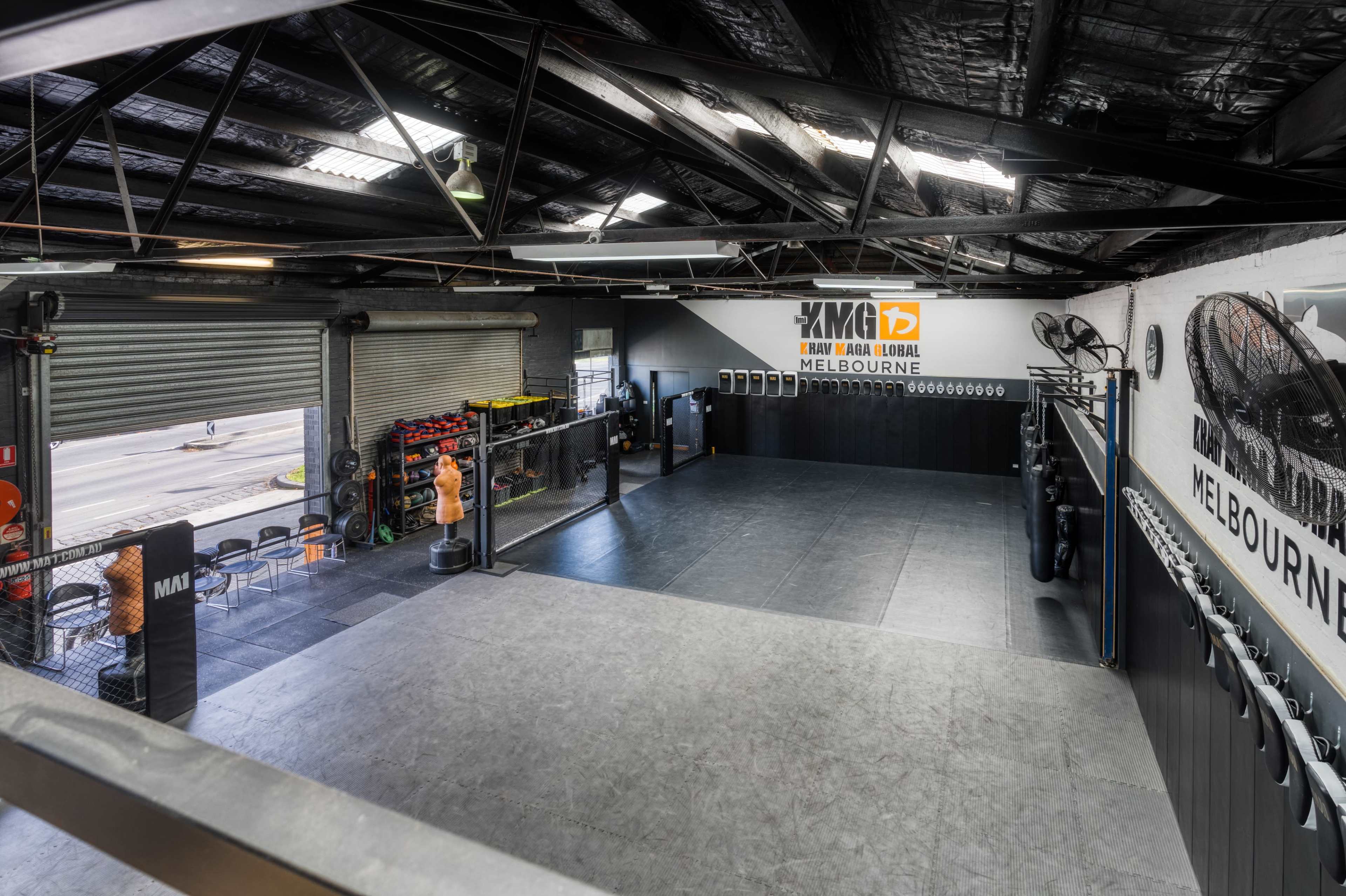 An indoor mixed martial arts training facility with an open training area, black mats, and gym equipment along the walls.