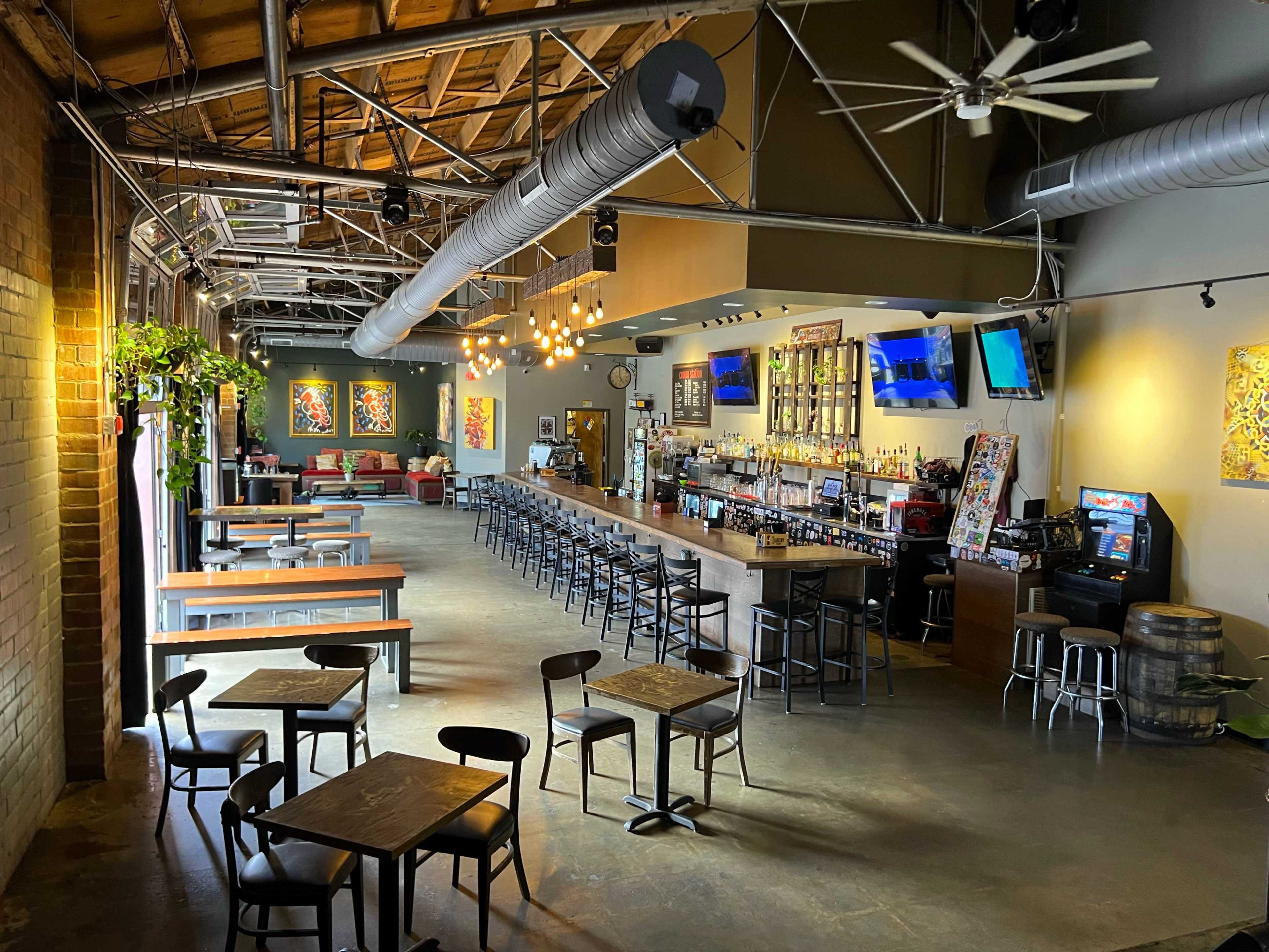 A spacious bar and dining area with a long wooden counter, rows of black stools, and tables scattered throughout, illuminated by pendant lights and adorned with colorful artwork on the walls.
