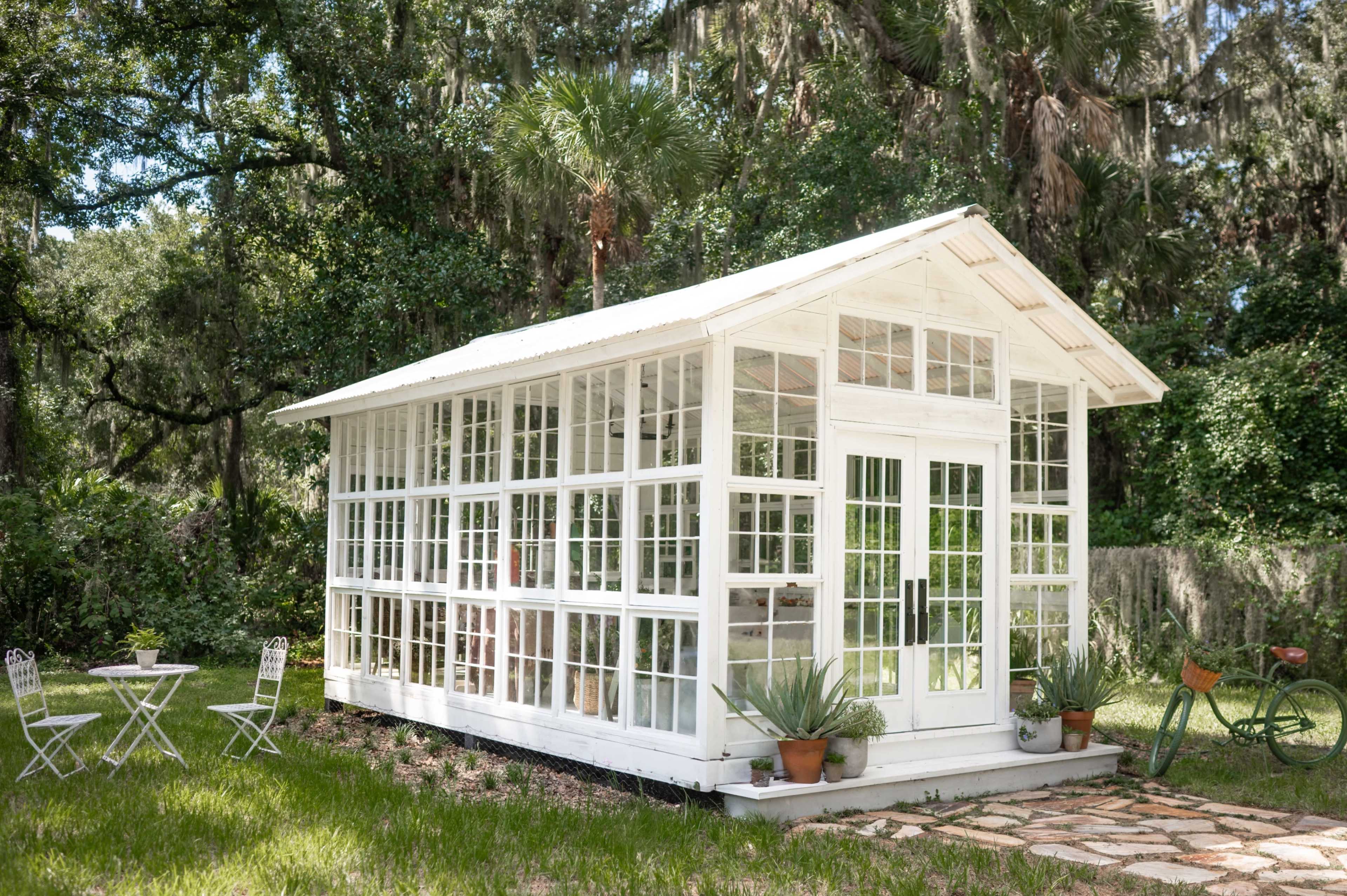 A white greenhouse with large glass panels is surrounded by greenery and features a small patio with two chairs and a bicycle nearby.