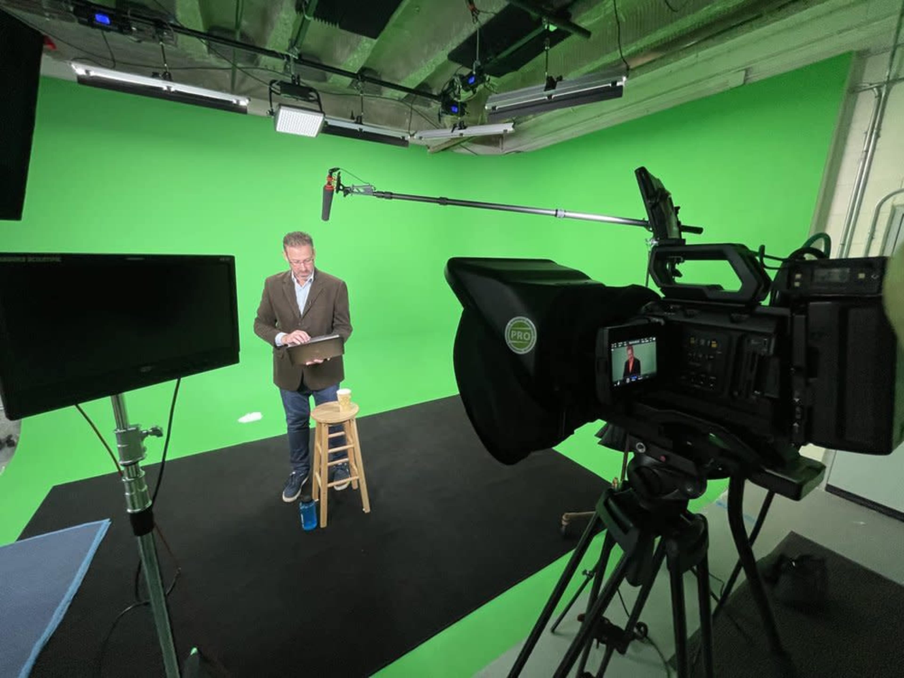 A person stands on a stool in front of a green screen, holding a tablet, with a camera and lighting equipment positioned nearby.