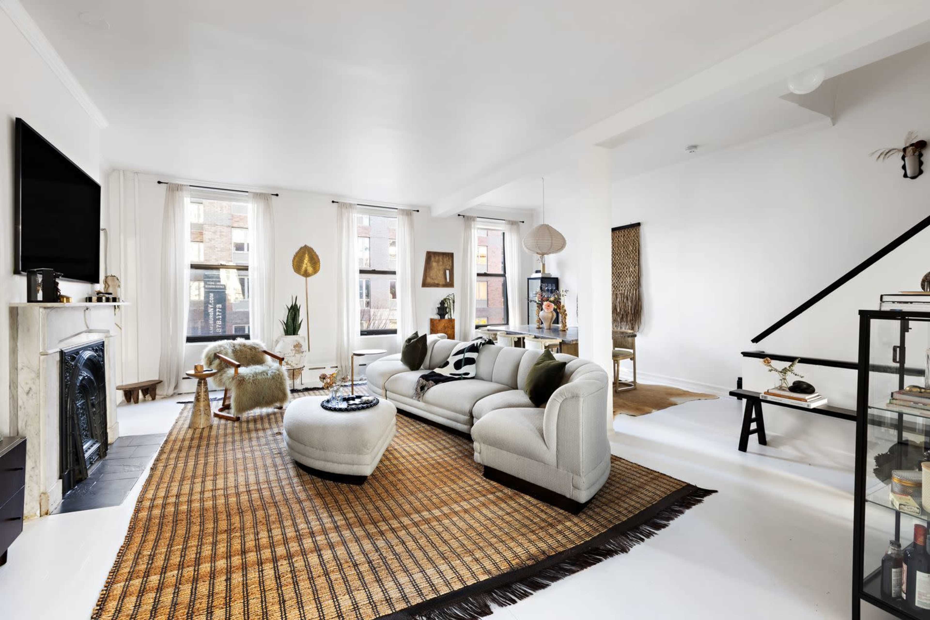 The image shows a modern living room with a gray sofa, a large woven rug, and multiple windows allowing natural light to fill the space.