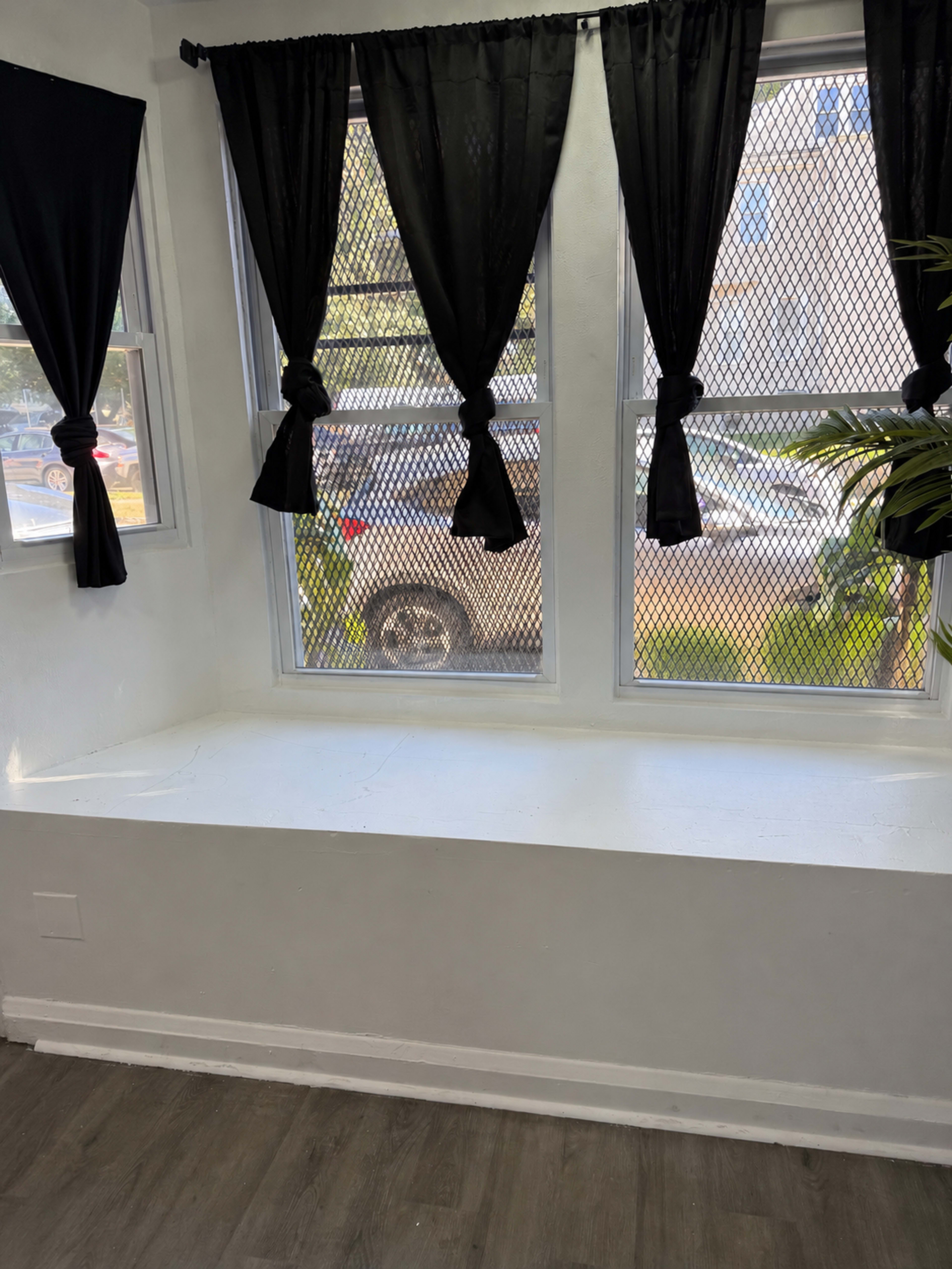 The image shows a sunlit room with a white window seat, two black curtains tied to the sides of the windows, and a view of a vehicle outside.