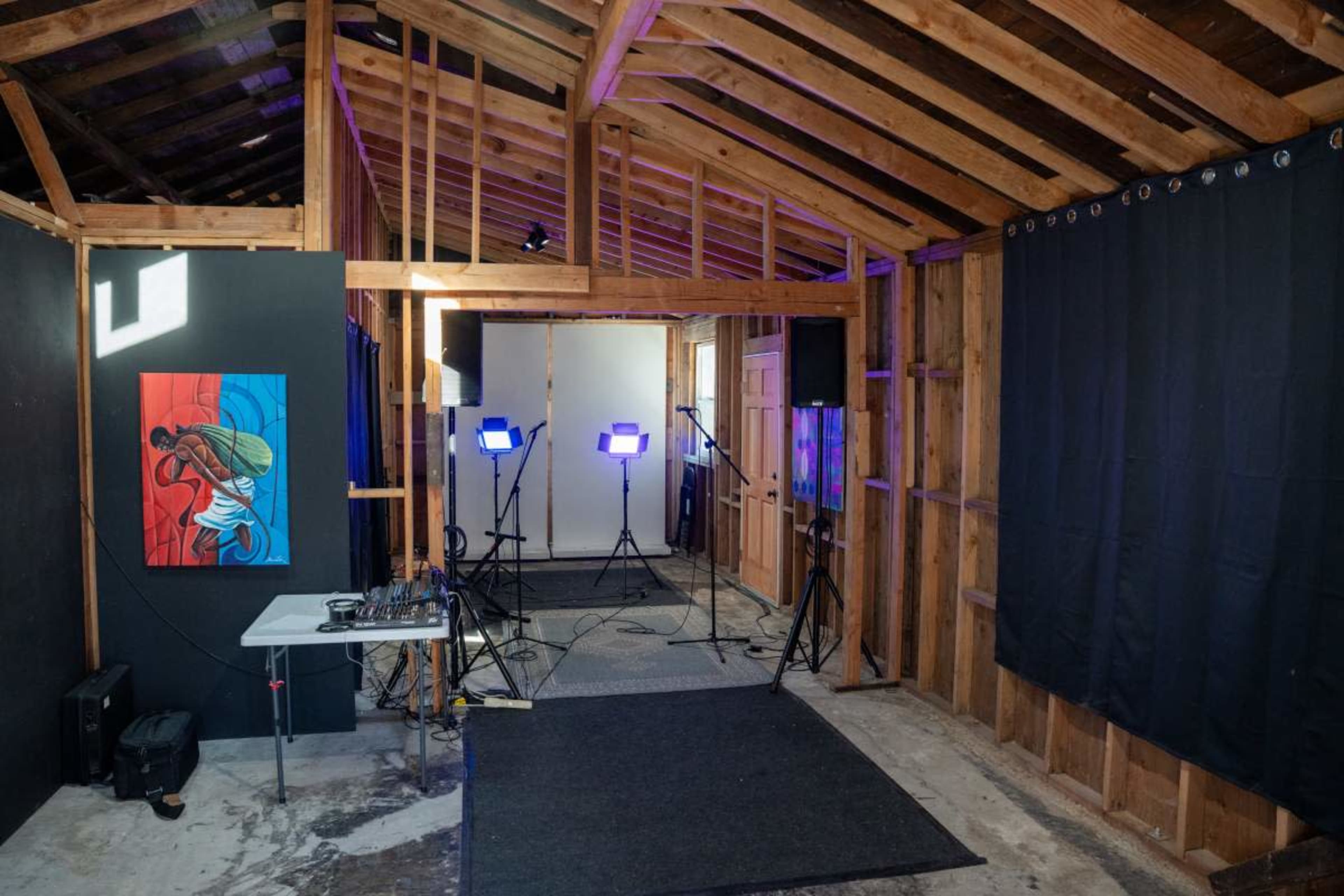 The image shows an unfinished interior of a wooden structure, featuring artwork on an adjacent wall, studio lights on tripods, and a table with equipment.