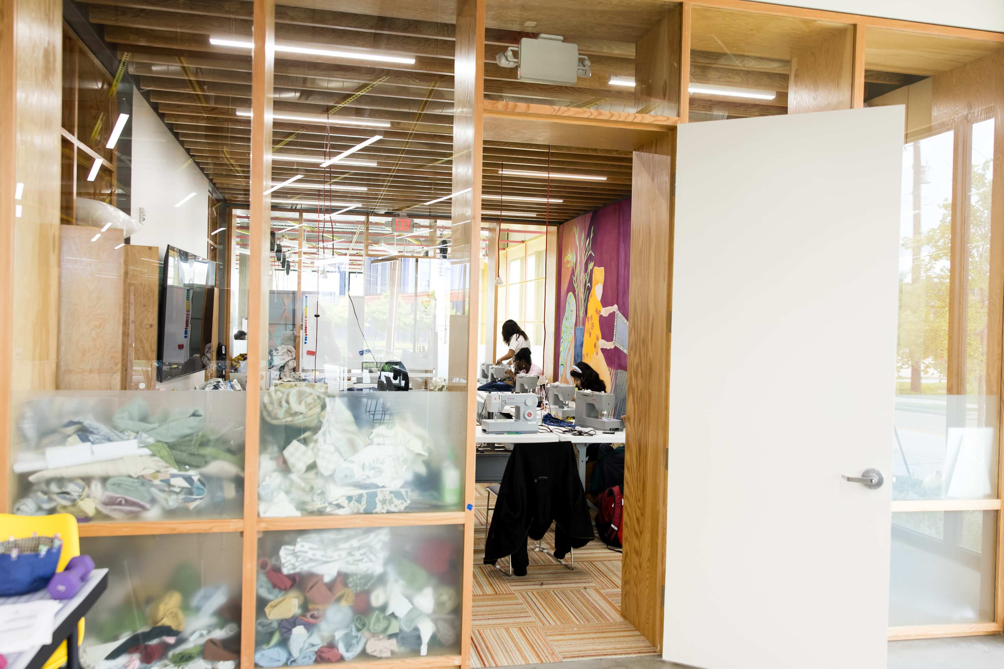 A bright workspace features large glass walls with several desks scattered inside, where people are engaged in various activities related to fabric and design.
