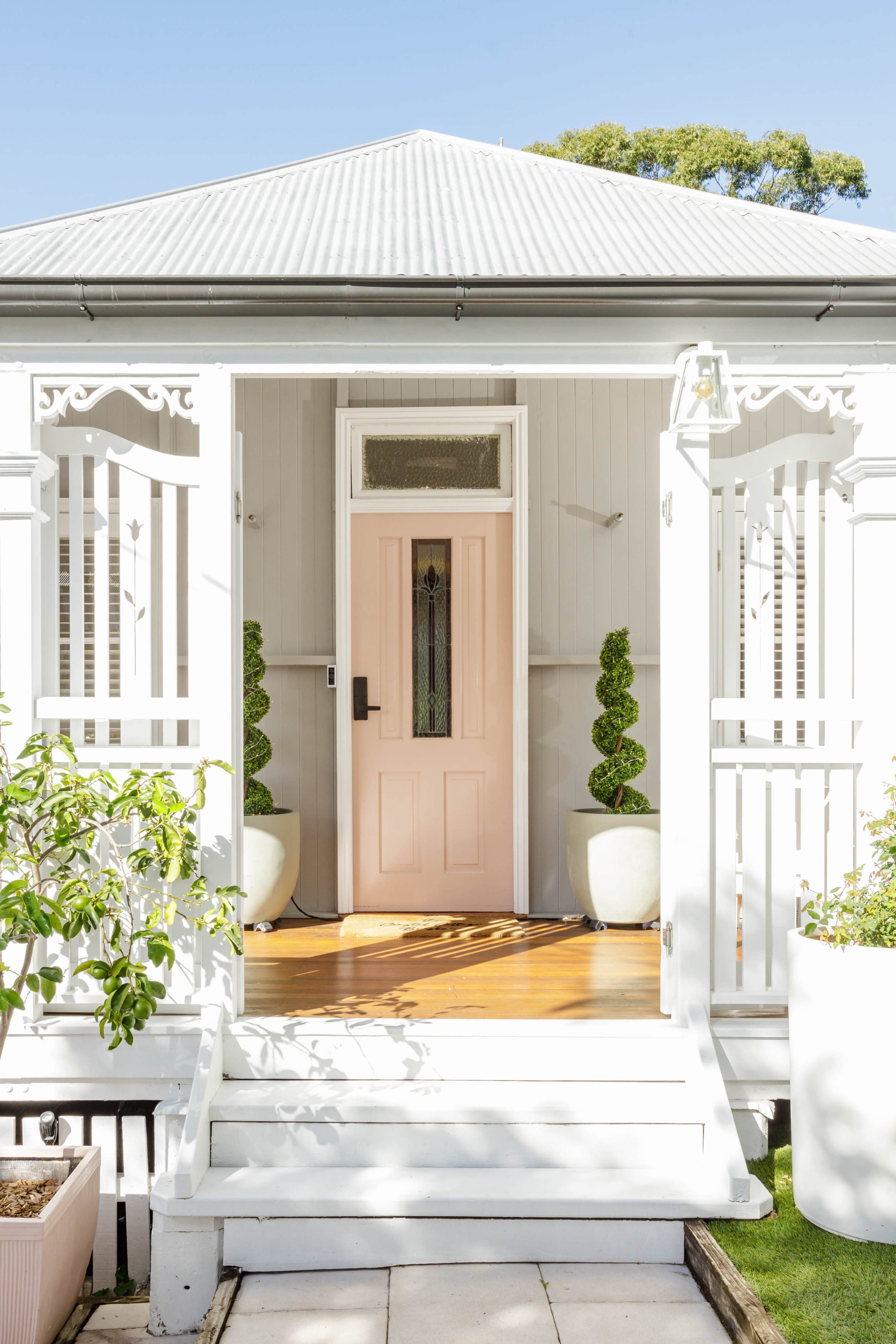 The image shows a front porch of a house with a pink door, surrounded by white railings and two potted topiary plants.
