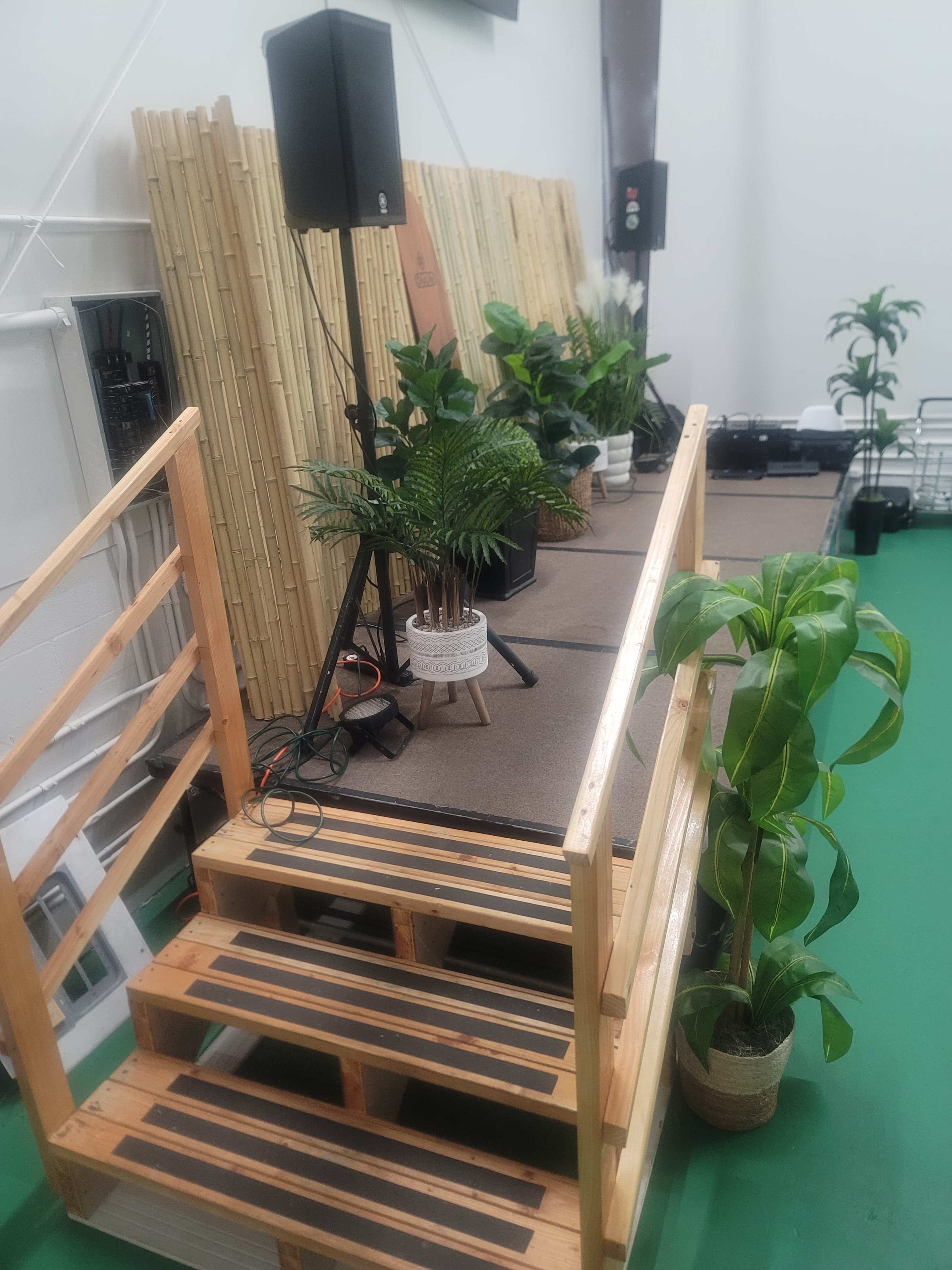 The image shows a small stage with wooden steps, decorated with potted plants and a bamboo backdrop, along with audio equipment on either side.