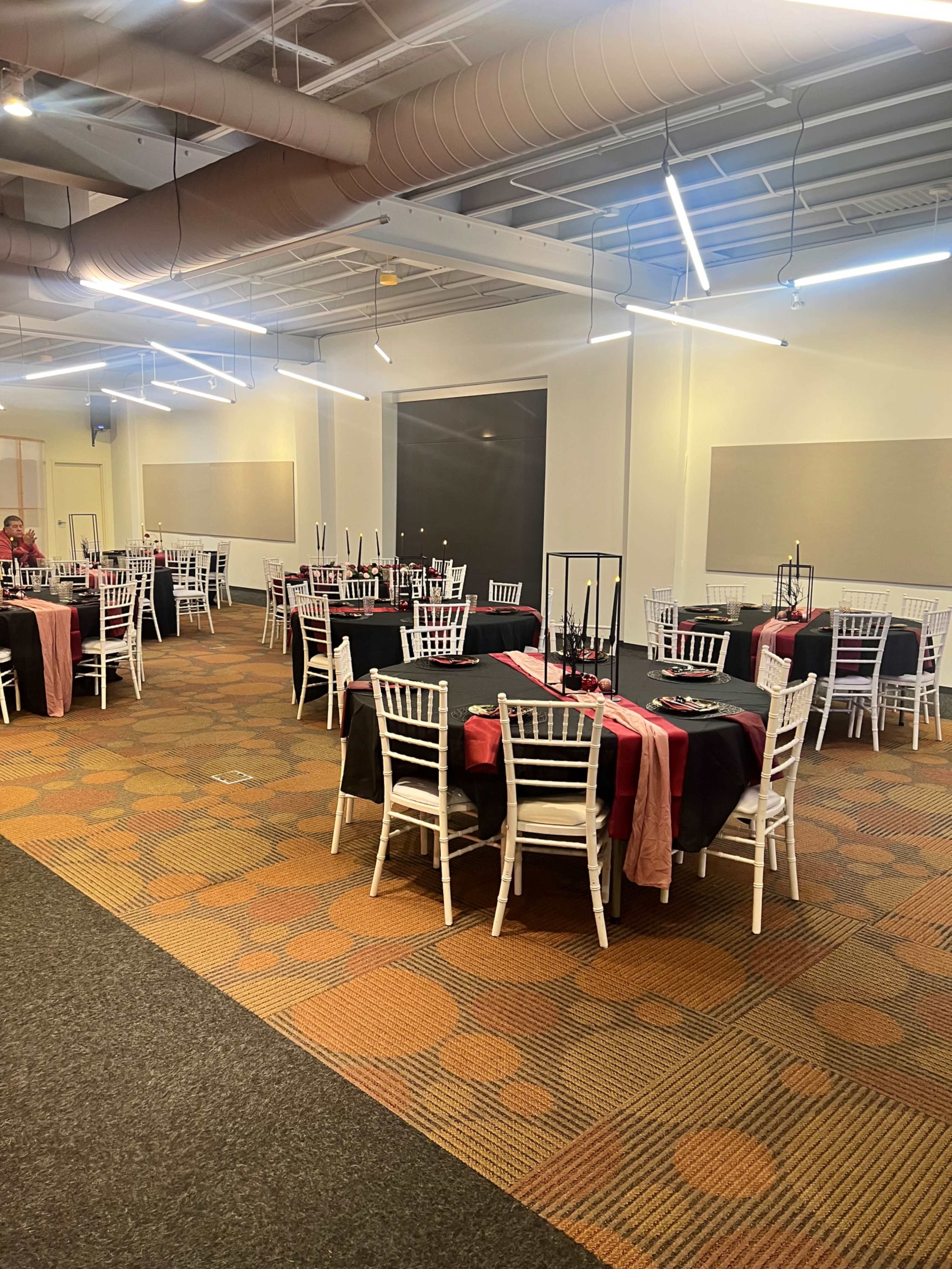 A spacious event room set with several round tables covered in black and red tablecloths, surrounded by white chairs.